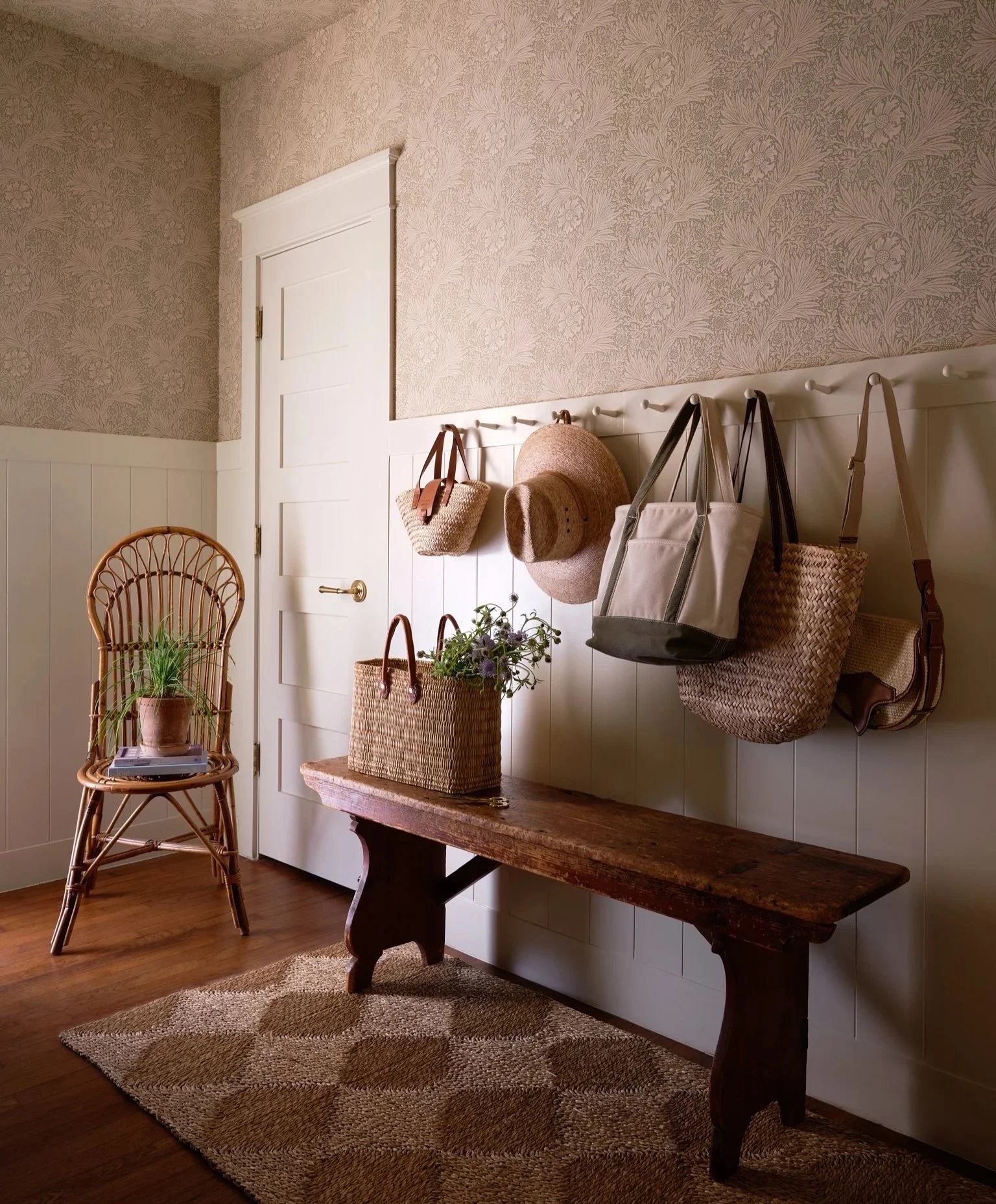 A cozy entryway with a rustic wooden bench, a woven basket, a potted plant, and a wall with hooks holding handbags and hats, a white door, and floral wallpaper.