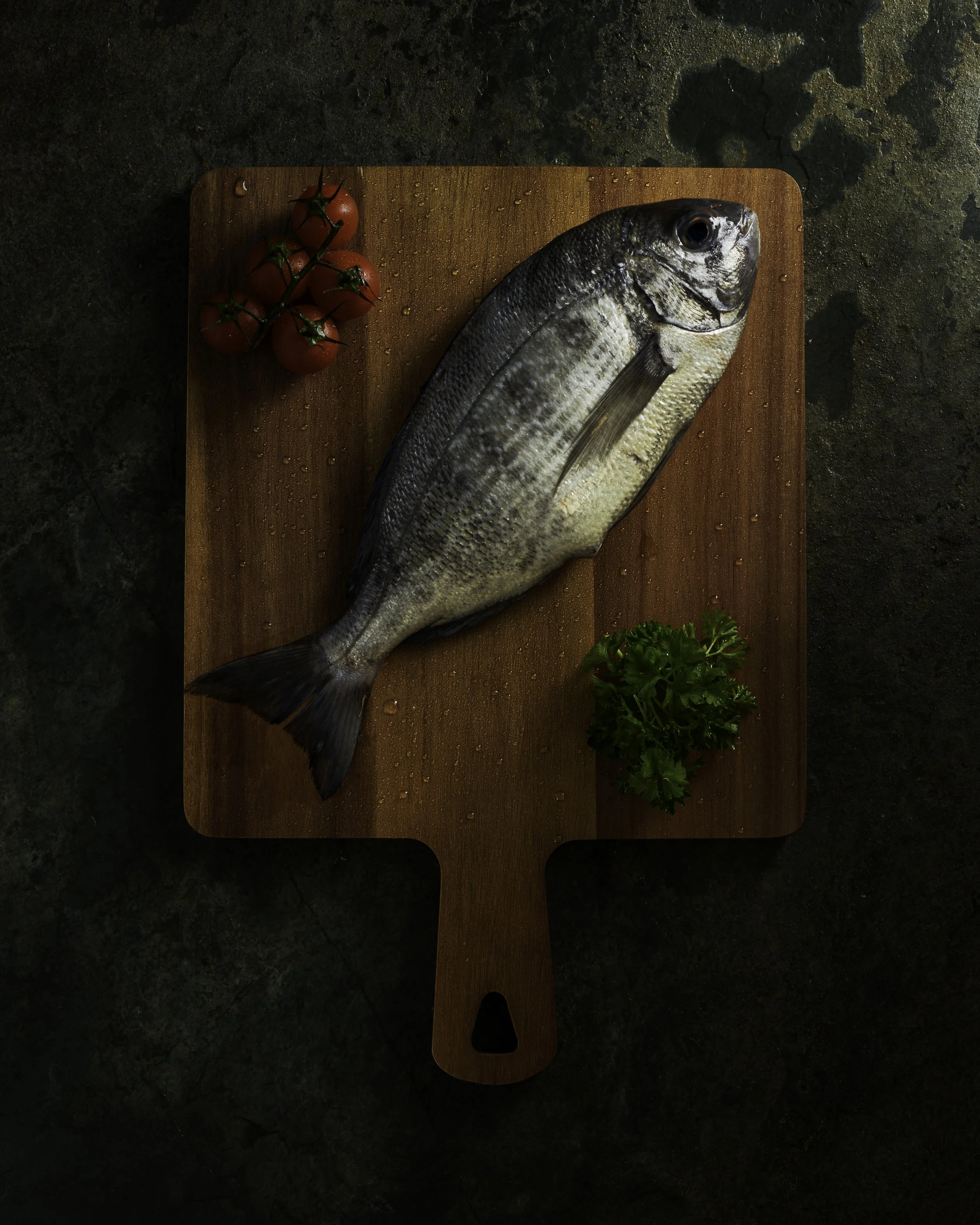 Dark and moody sea food photography created by commercial photographer at Emily Mills Photographic Studio. Sea bream on wooden board with fresh tomatoes and garnish. board is placed on a slate background https://www.emilymillsphotography.com/foodandd