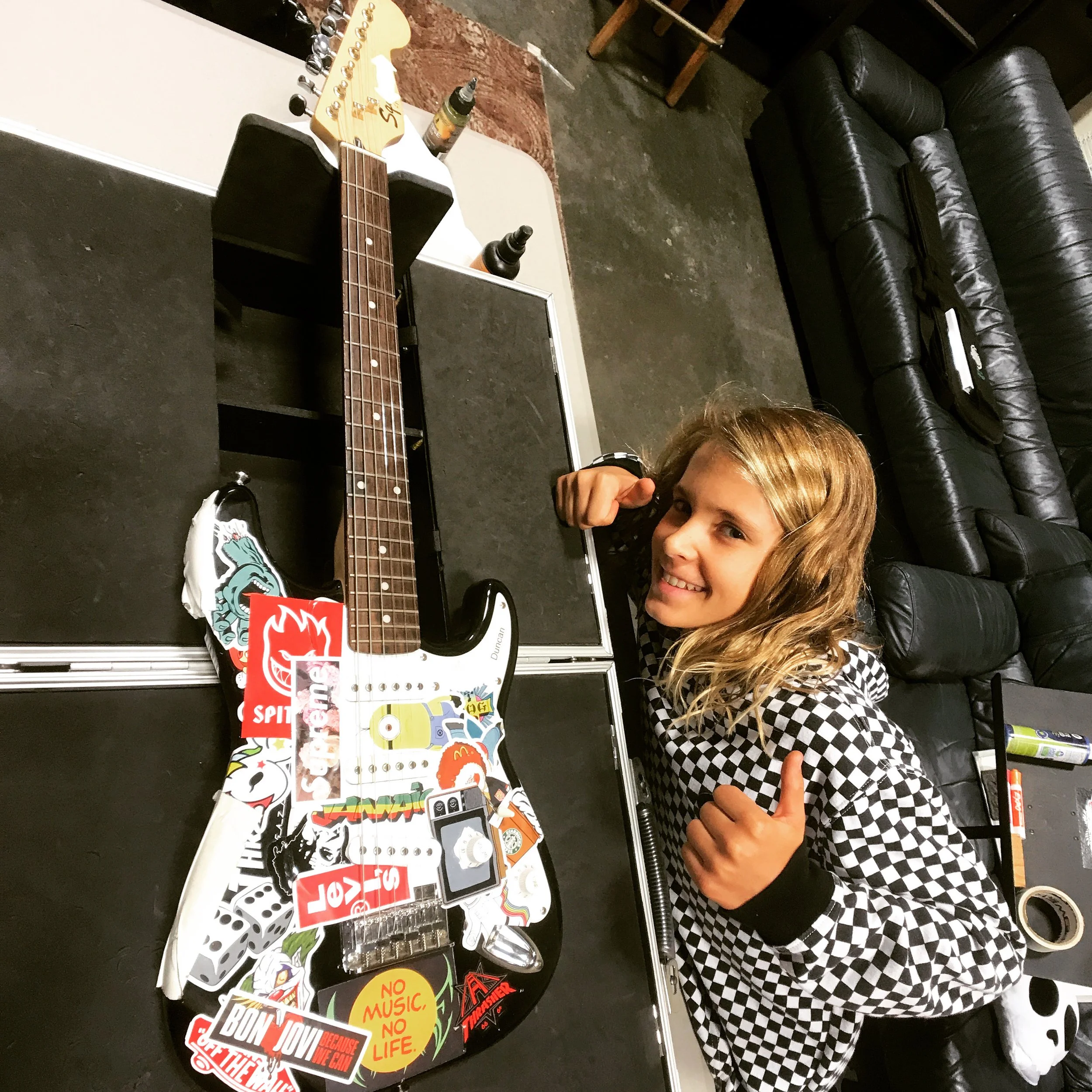 A young girl with wavy blonde hair, wearing a black and white checkered hoodie, gives a thumbs-up while sitting at a table with a guitar covered in colorful stickers. The stickers include brands, logos, and phrases like "Supreme," "No Music, No Life," and "Levi's." The background features black leather couches and a concrete floor.