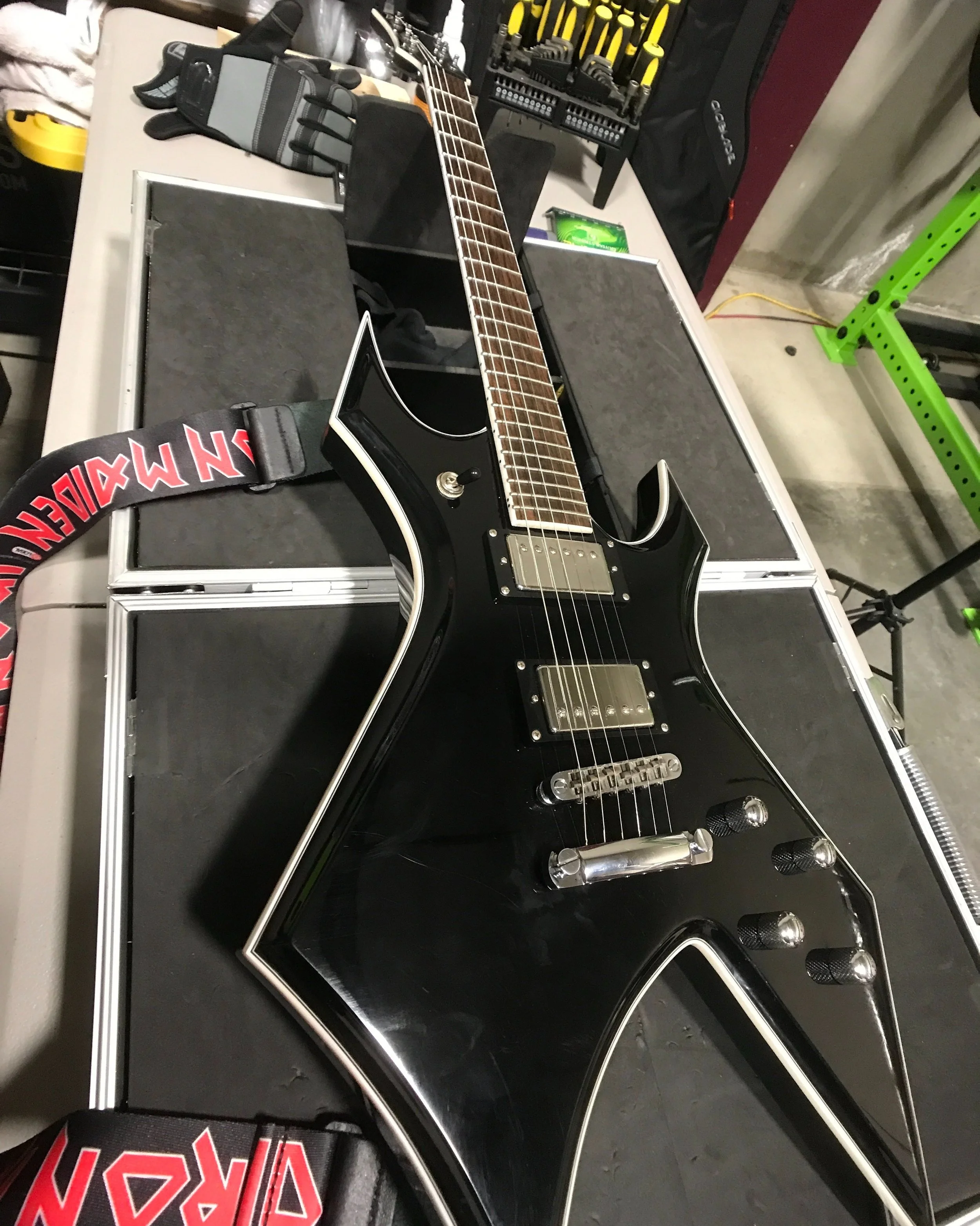Black electric guitar with white trim and chrome hardware resting on a flight case, surrounded by tools and equipment in a workshop or studio setting.