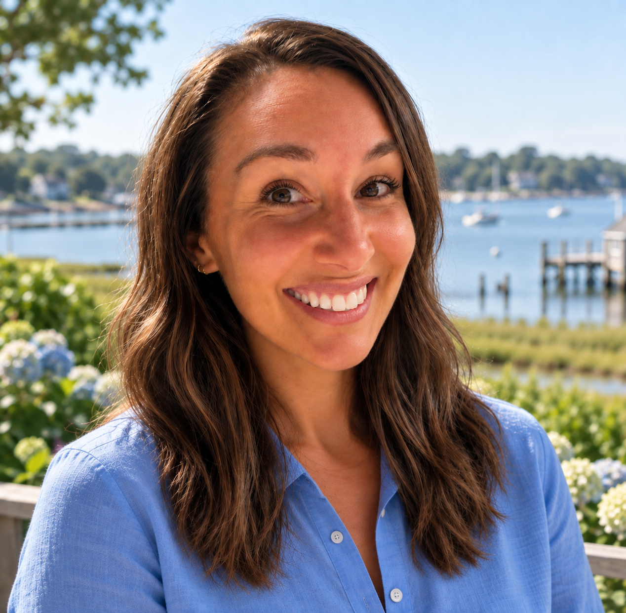 A smiling woman with dark hair tied back, wearing a light denim jacket and a gray t-shirt, standing outdoors in front of a wooden fence and greenery.