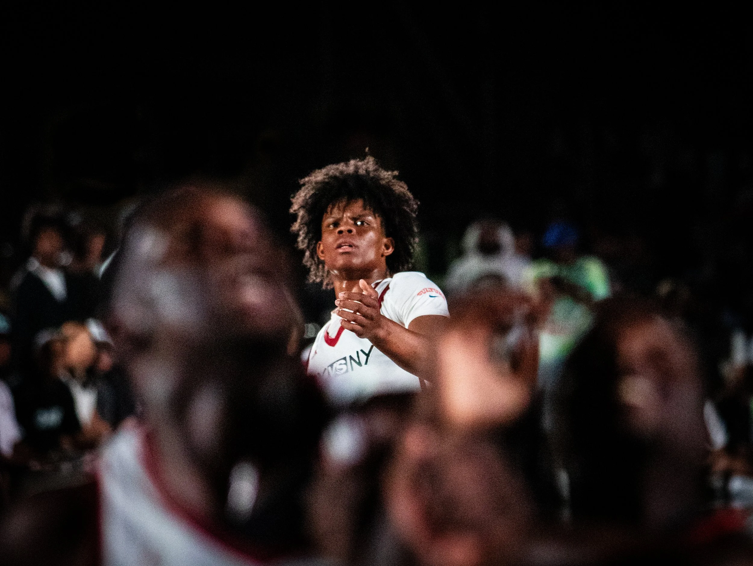 A young woman with curly hair wearing a white sports jersey stands among a crowd at night, looking concerned or surprised.