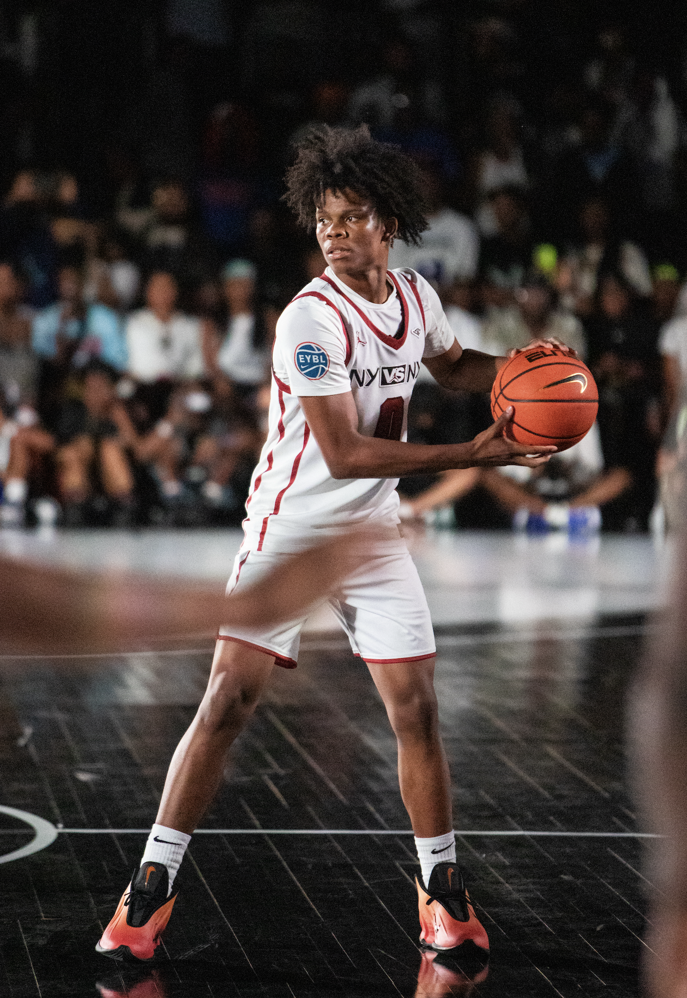 A female basketball player with athletic build in a white and red uniform holding an orange basketball on a dark indoor court, with a crowd in the background.