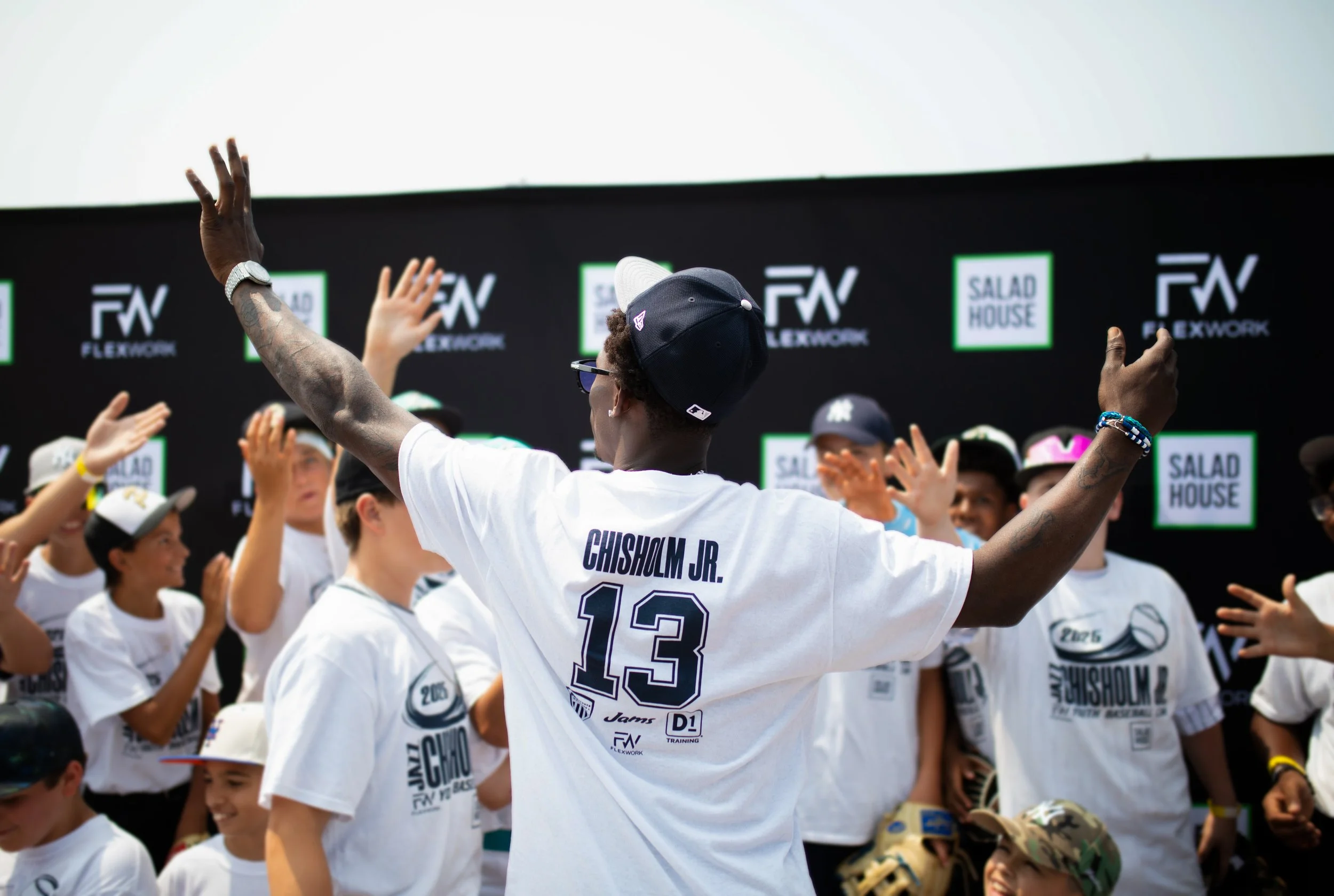 A group of children and a man celebrating outdoors, with the man wearing a white jersey with the name Chisholm Jr. and the number 13 on the back. They are raising their hands, and the background features black and green signs with the words 'Flexwork