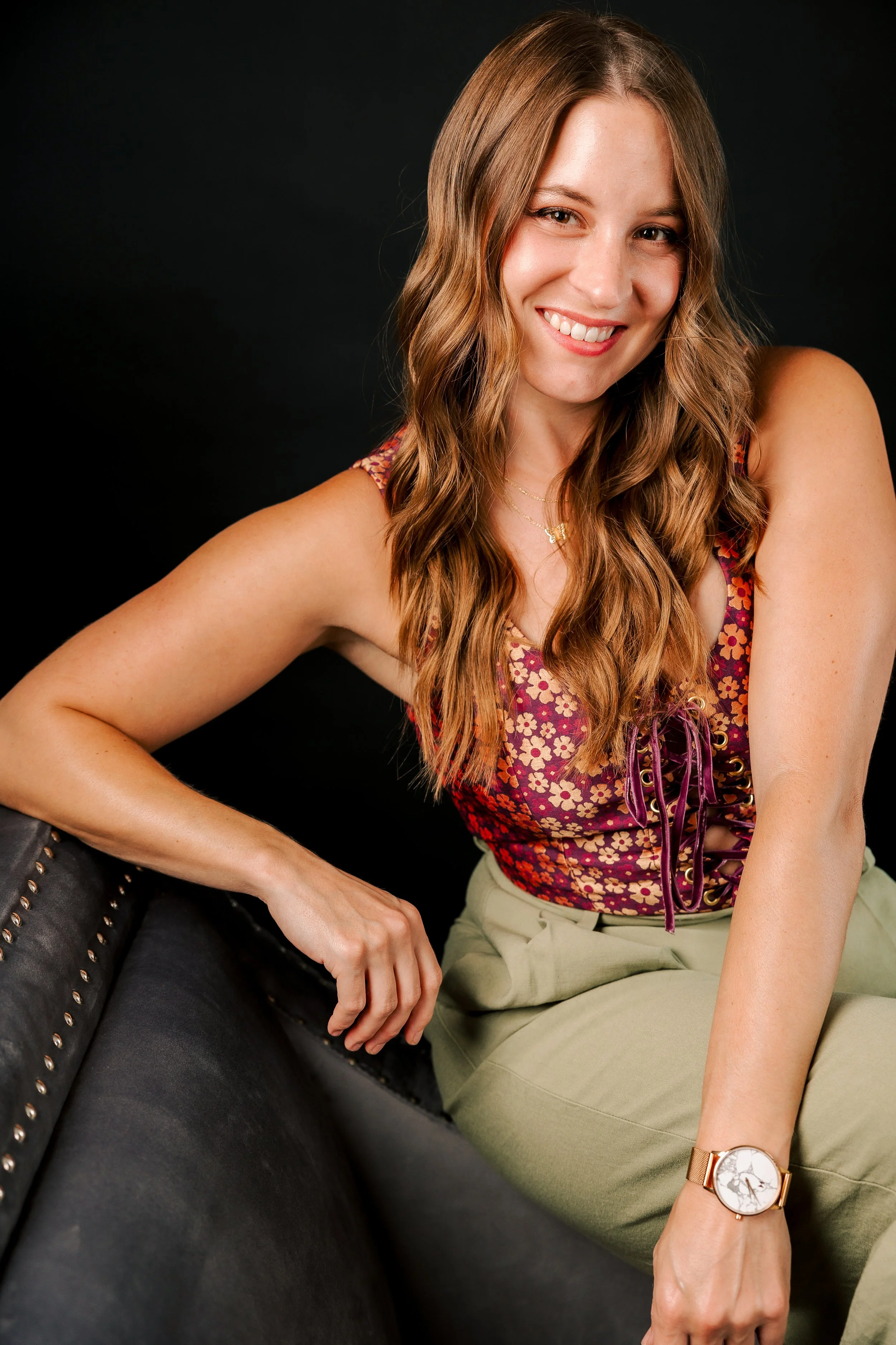 A woman with long, styled brown hair smiling, wearing a floral sleeveless top with lace-up detail, beige pants, and a watch, sitting on a black leather chair against a black background.
