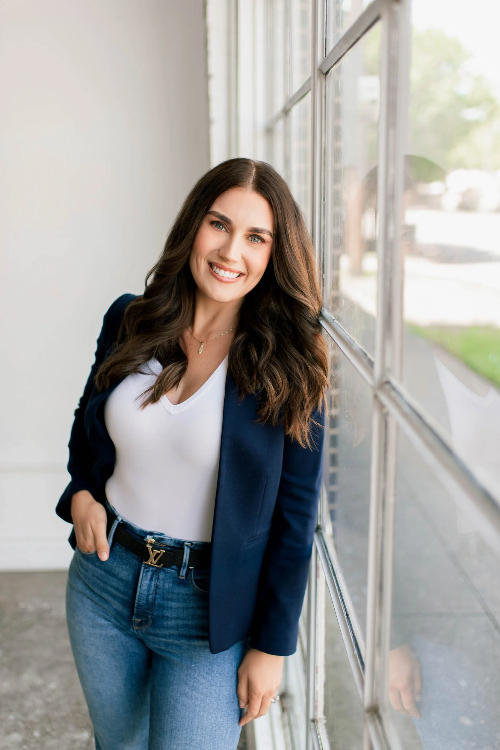 A woman with long brown hair and light skin, smiling, standing by a large window with a city view, wearing a navy blazer, white top, and blue jeans. headshots in Frisco TX, branding photography DFW, Prosper TX headshots, branding photography Plano TX