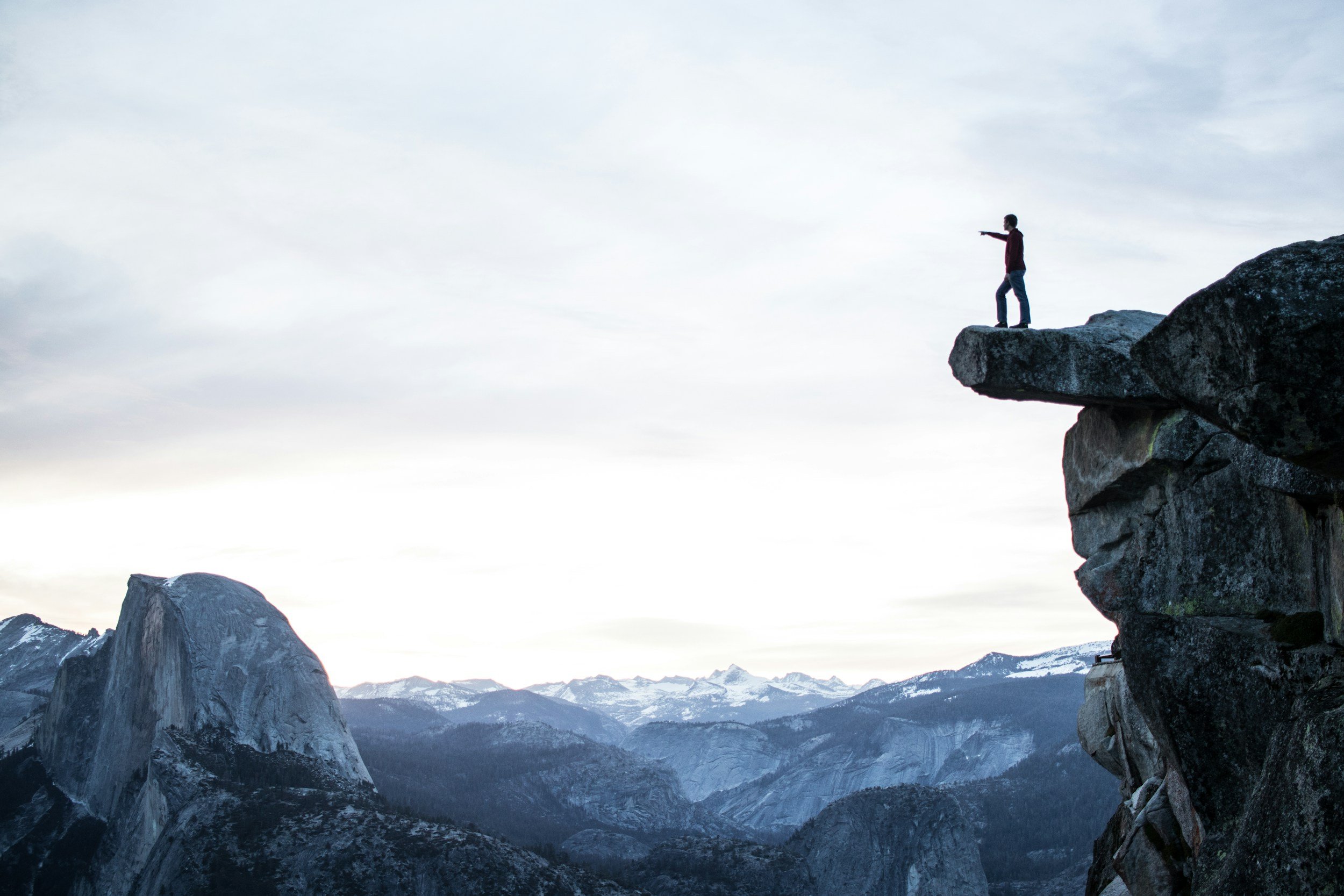 A person standing on the edge of a rocky cliff, pointing into the distance, with a mountain landscape and sky in the background.