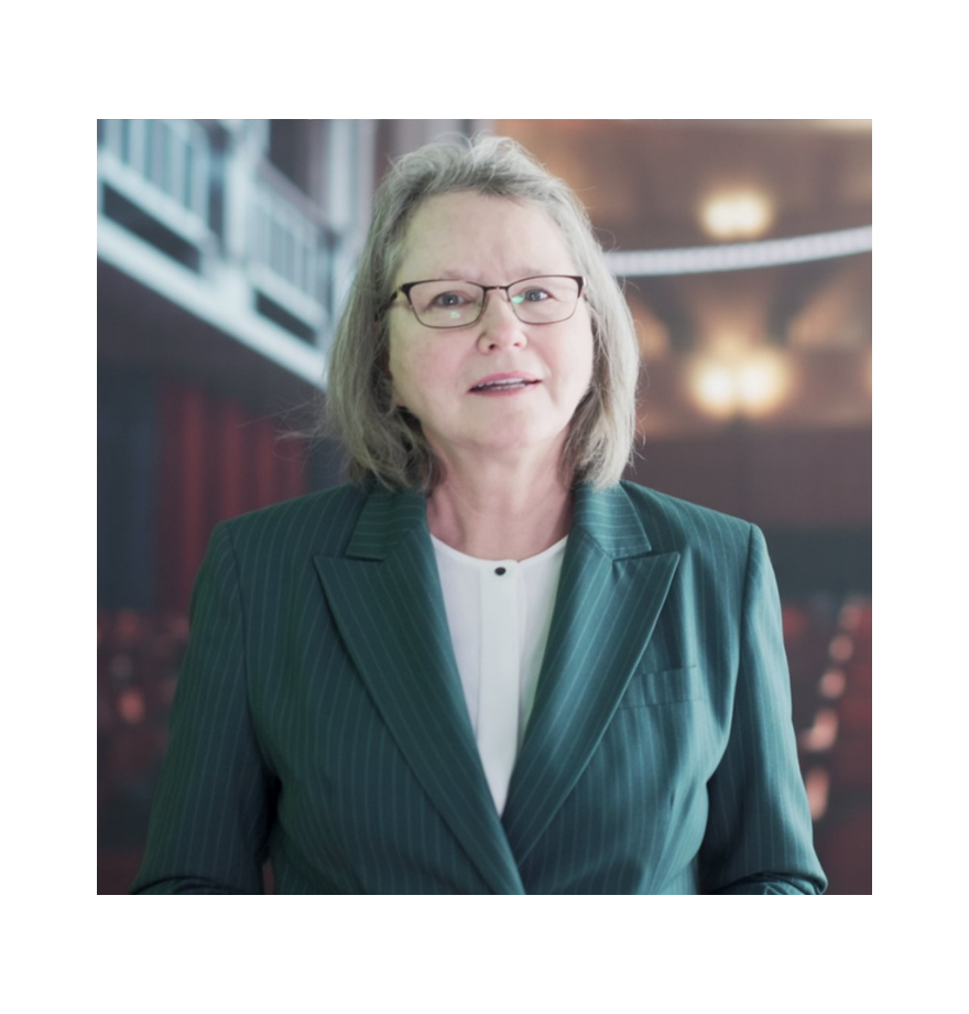 A middle-aged woman with gray hair and glasses, wearing a dark green pinstripe blazer and a white blouse, standing in an indoor setting that resembles a theater or auditorium.