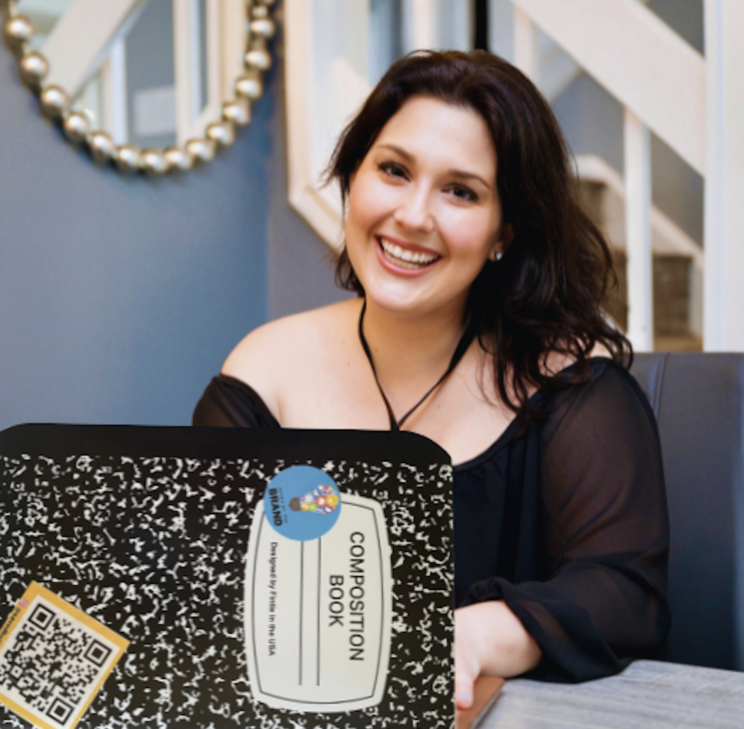 A woman with dark hair, wearing a black off-shoulder top, sitting at a table with a composition book labeled 'composition book' in front of her, smiling at the camera.