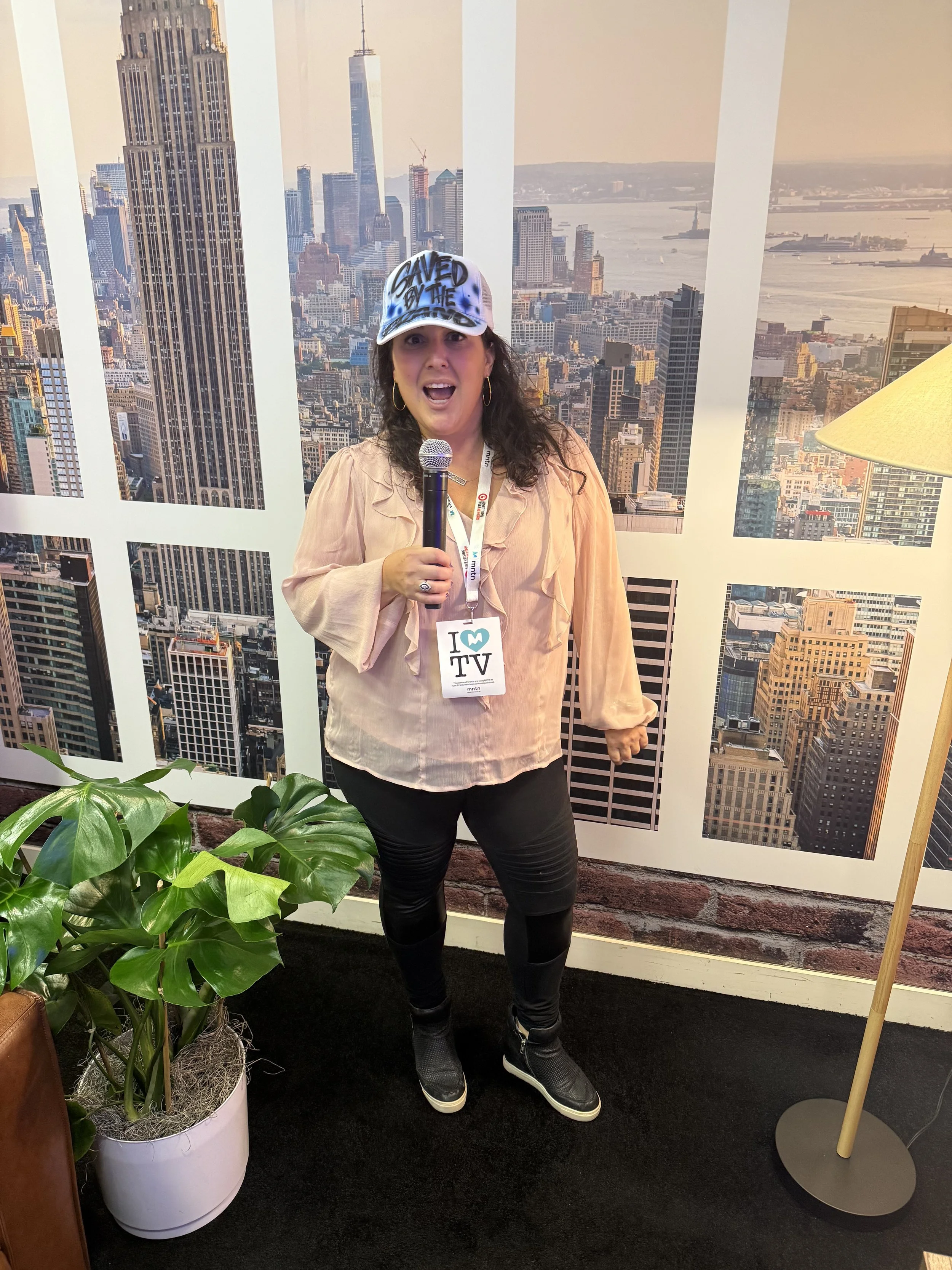 A woman standing in front of a wall with a photo of New York City skyline, holding a microphone, wearing a blue and white cap with 'Saved by the TV' written on it and a badge that says 'I ❤️ TV' around her neck.