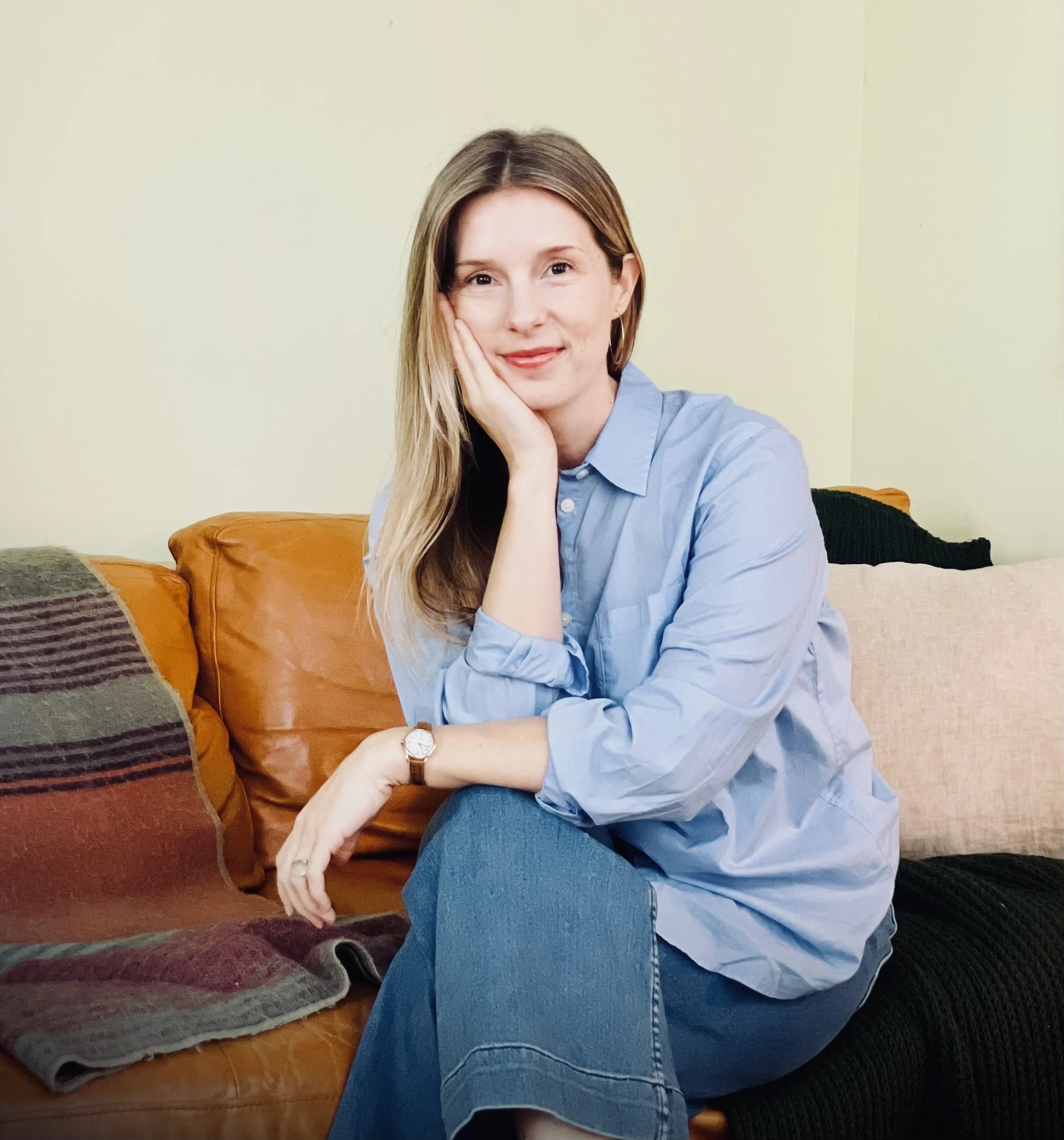 A woman with long, light brown hair and fair skin sitting on a sofa, wearing a light blue button-up shirt and blue jeans, smiling with her head resting on her left hand.