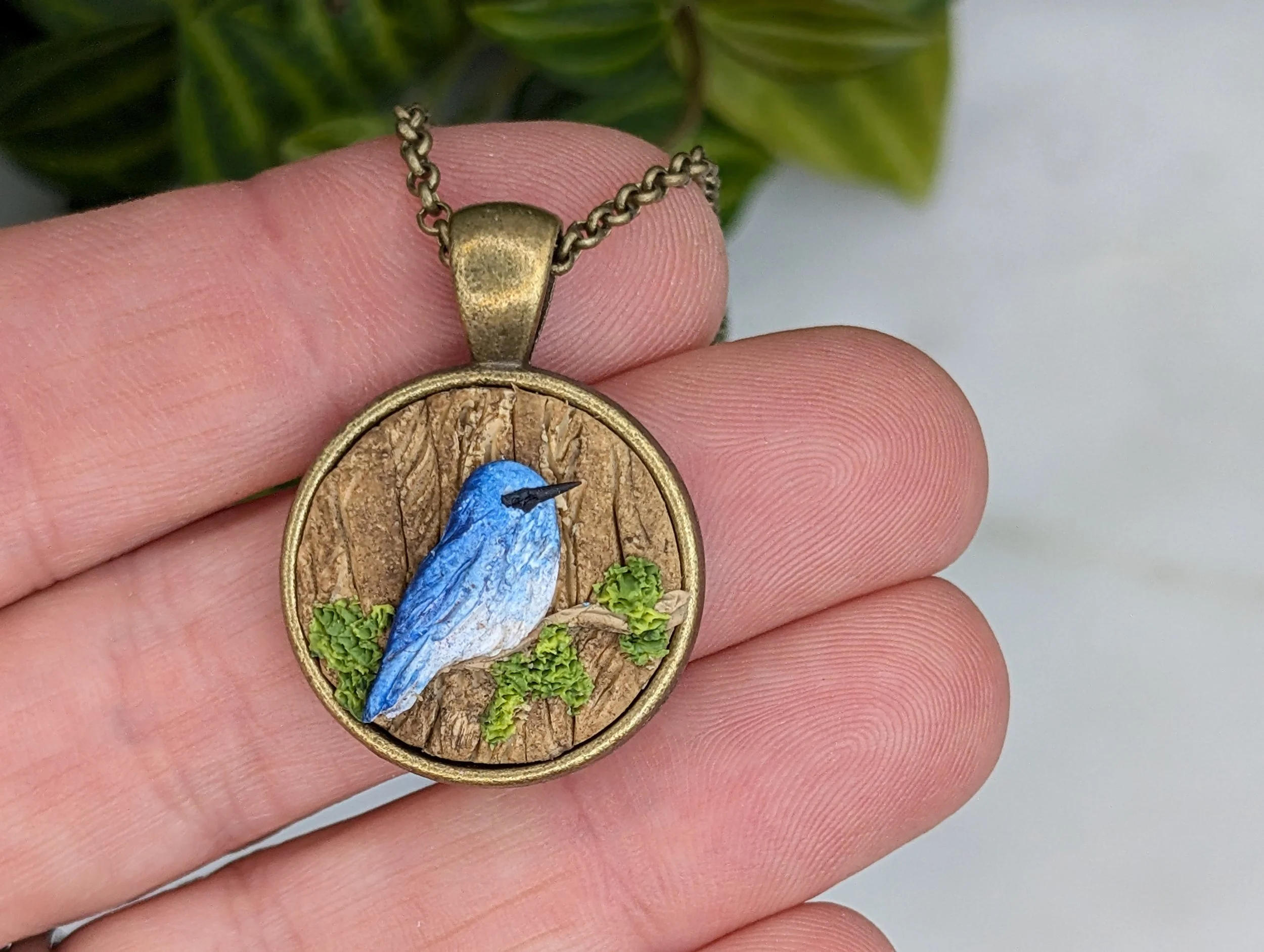 Handmade polymer clay and antique bronze pendant necklace featuring a tiny detailed mountain bluebird next to a textured, mossy tree trunk. By Woodsmoke and Willow.