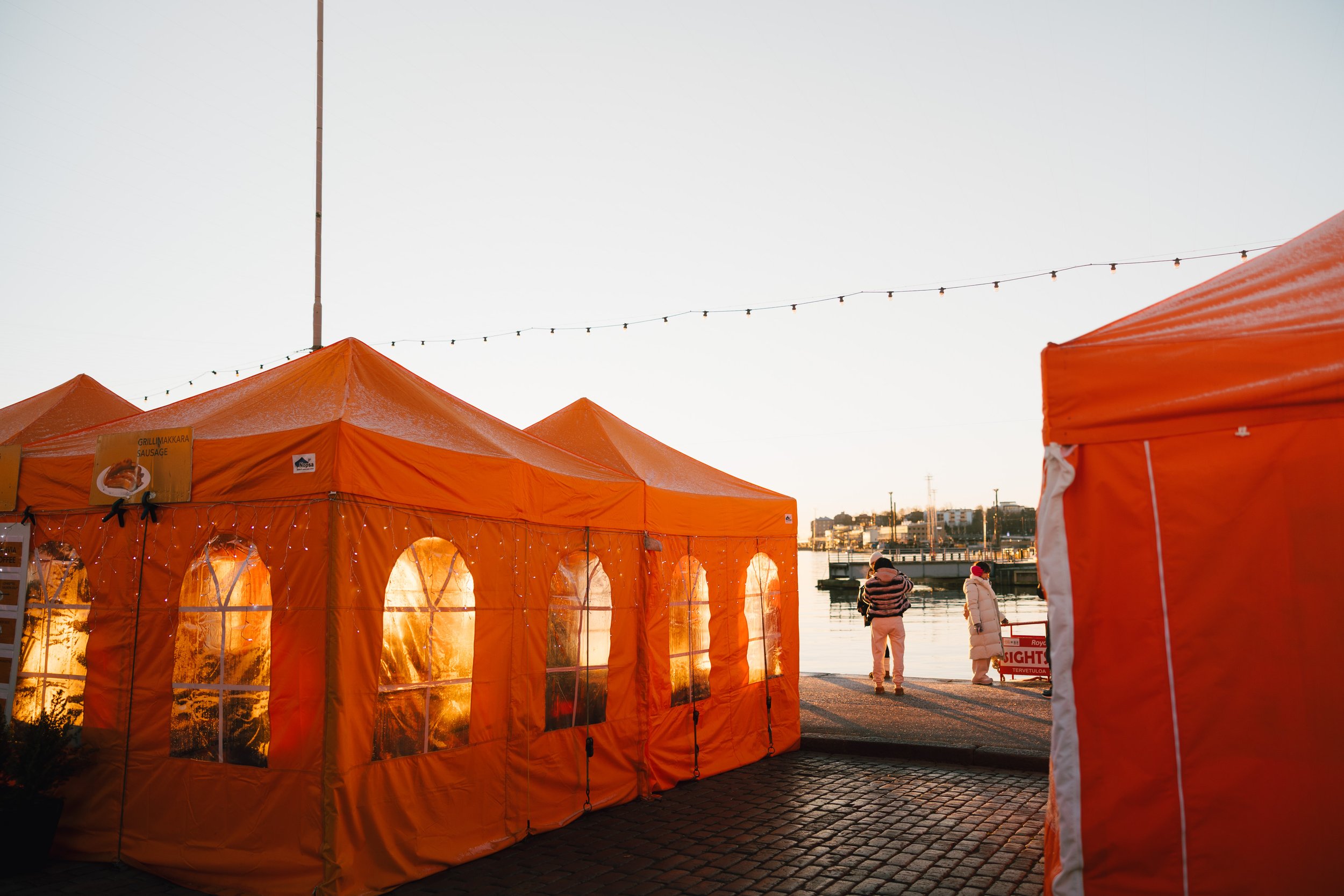 Helsinki Market Square, Orange tents set up near a waterfront during sunrise, with people walking in the background and string lights overhead.