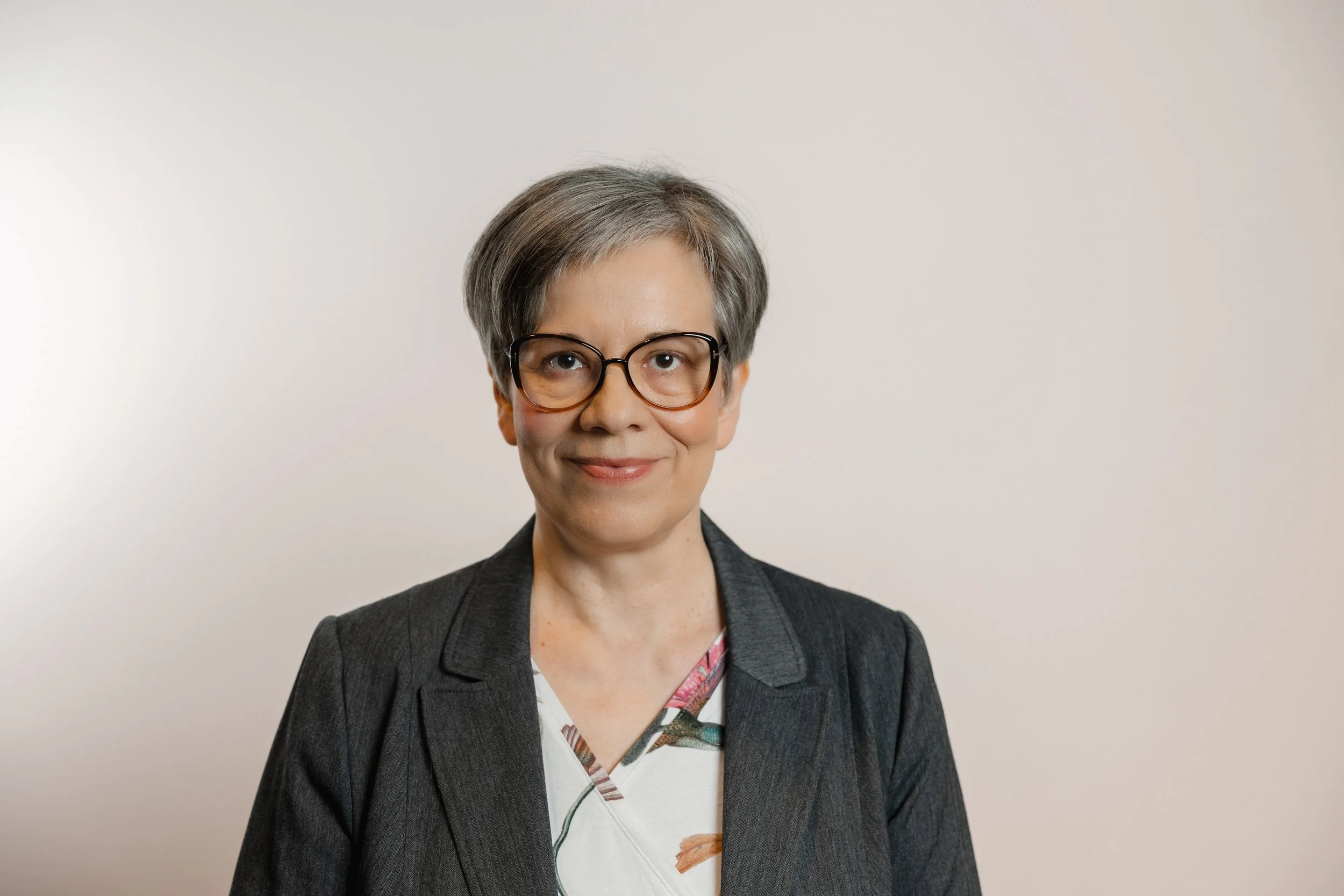 A woman with short gray hair, wearing glasses, a dark gray blazer, and a patterned blouse, smiling in front of a plain light-colored background.