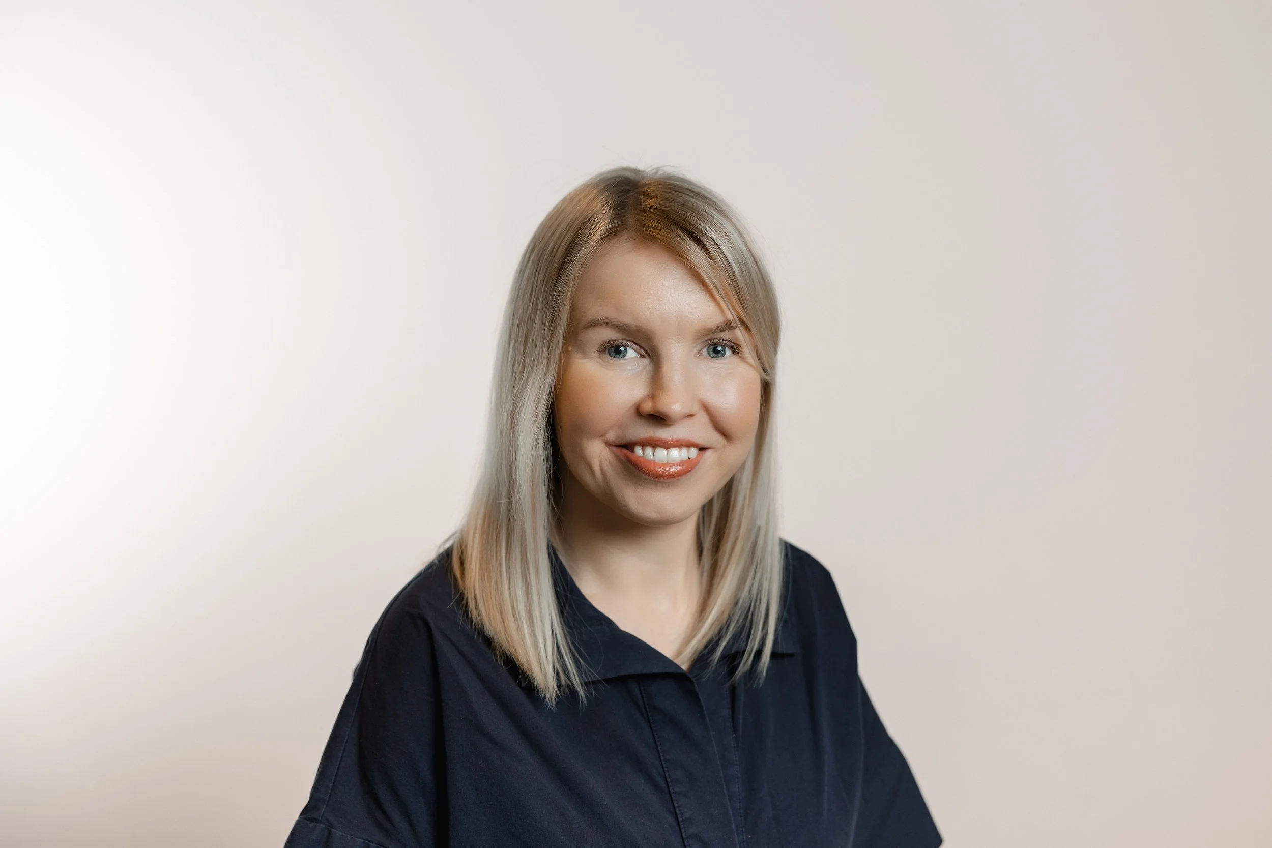 A woman with blonde hair and blue eyes smiling, wearing a navy blue shirt, against a plain light background.