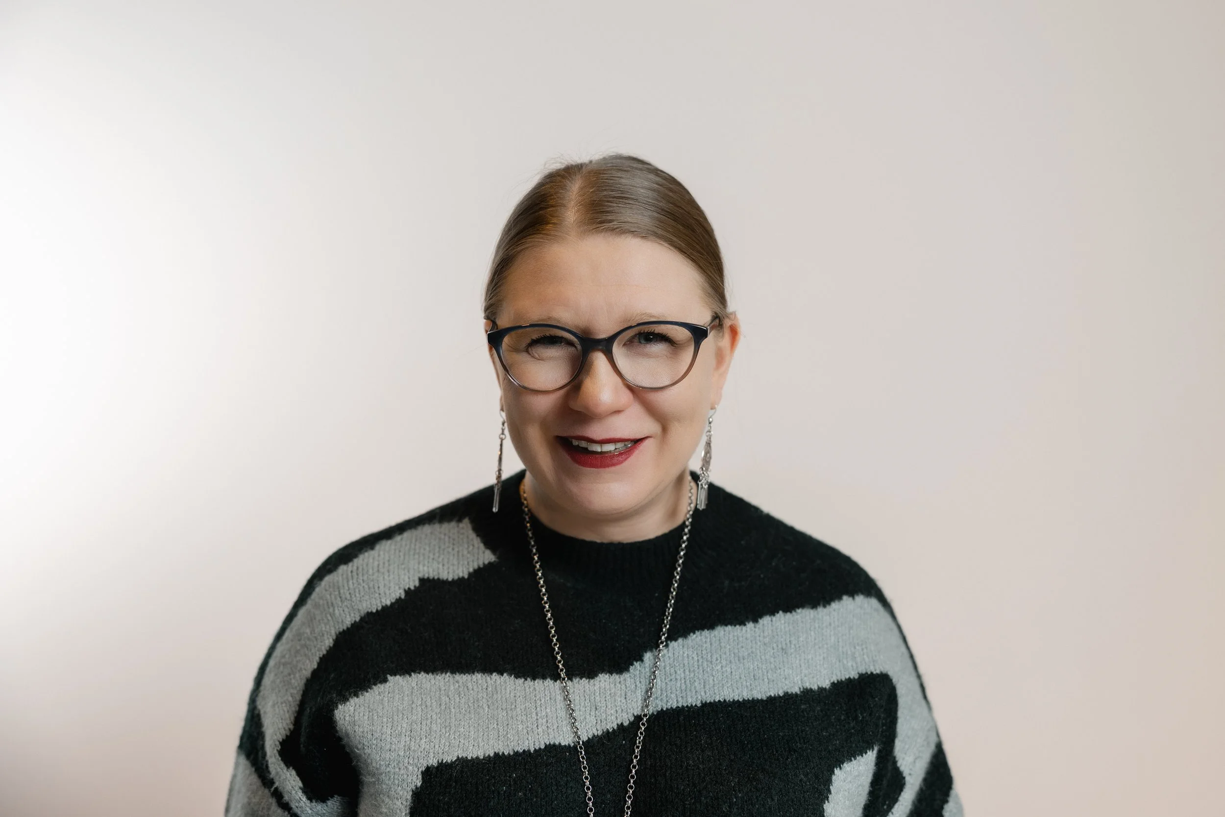 A woman with glasses, earrings, and a necklace smiling at the camera against a plain white background.