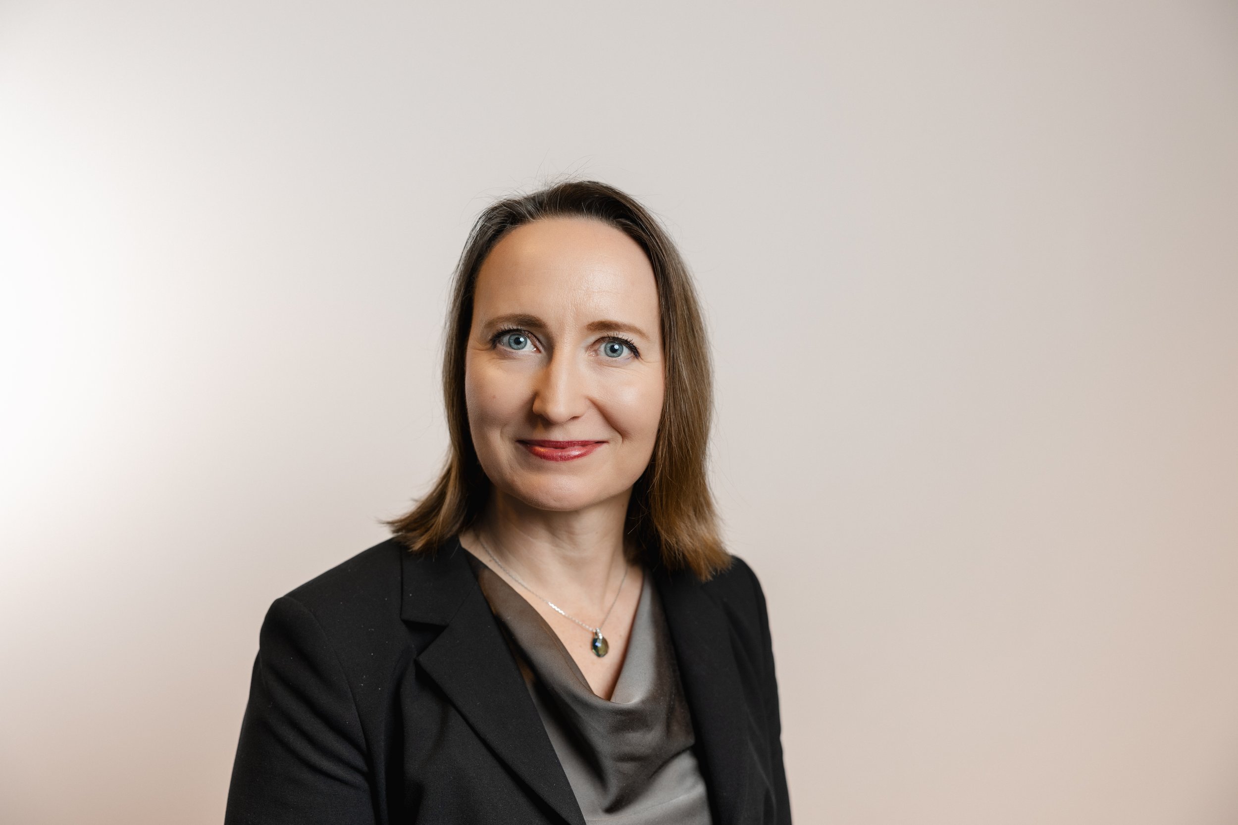 A woman with shoulder-length brown hair and blue eyes, wearing a black blazer and a gray blouse, smiling slightly in front of a plain white wall.