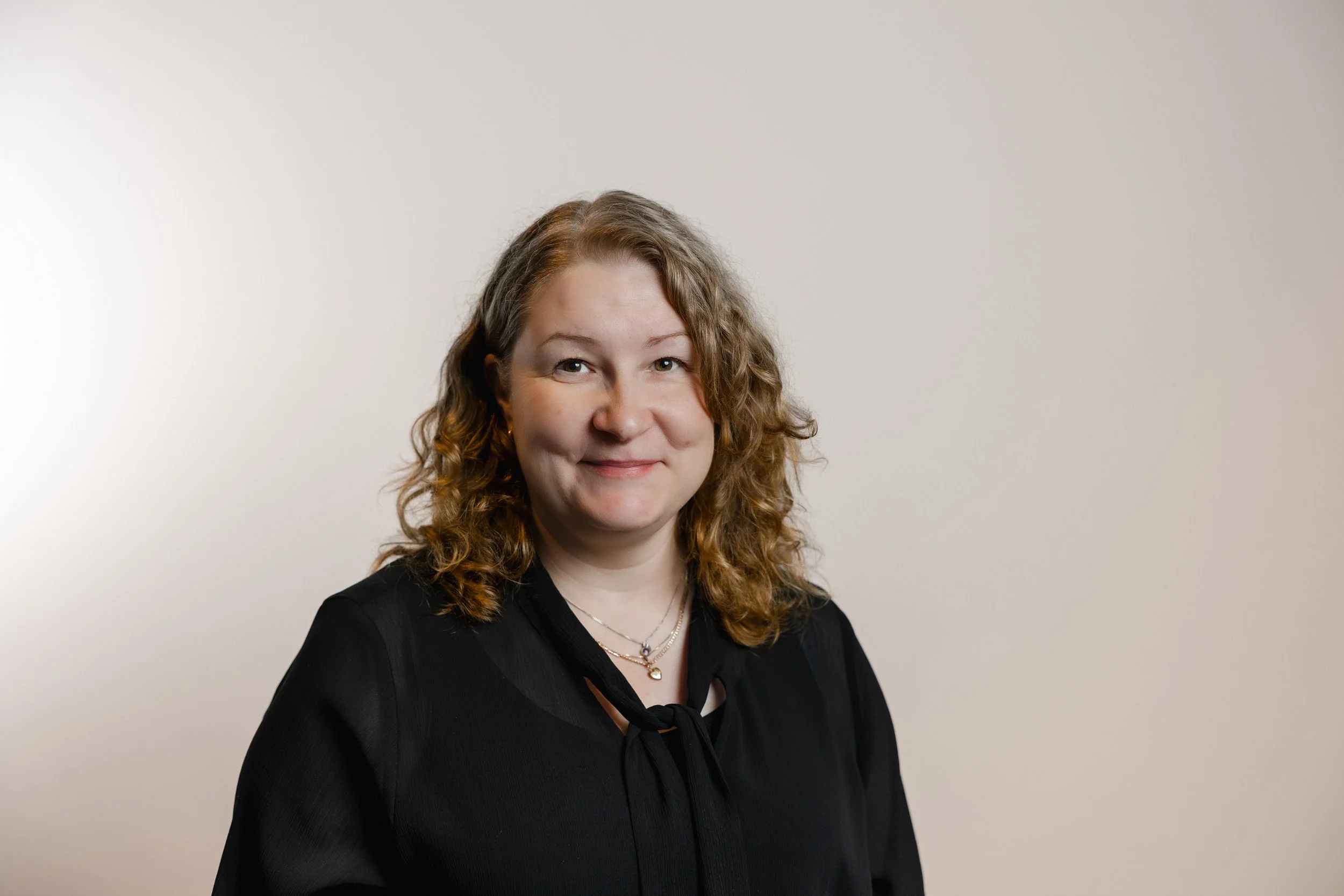 A woman with curly brown hair and a black blouse smiling in front of a plain background.