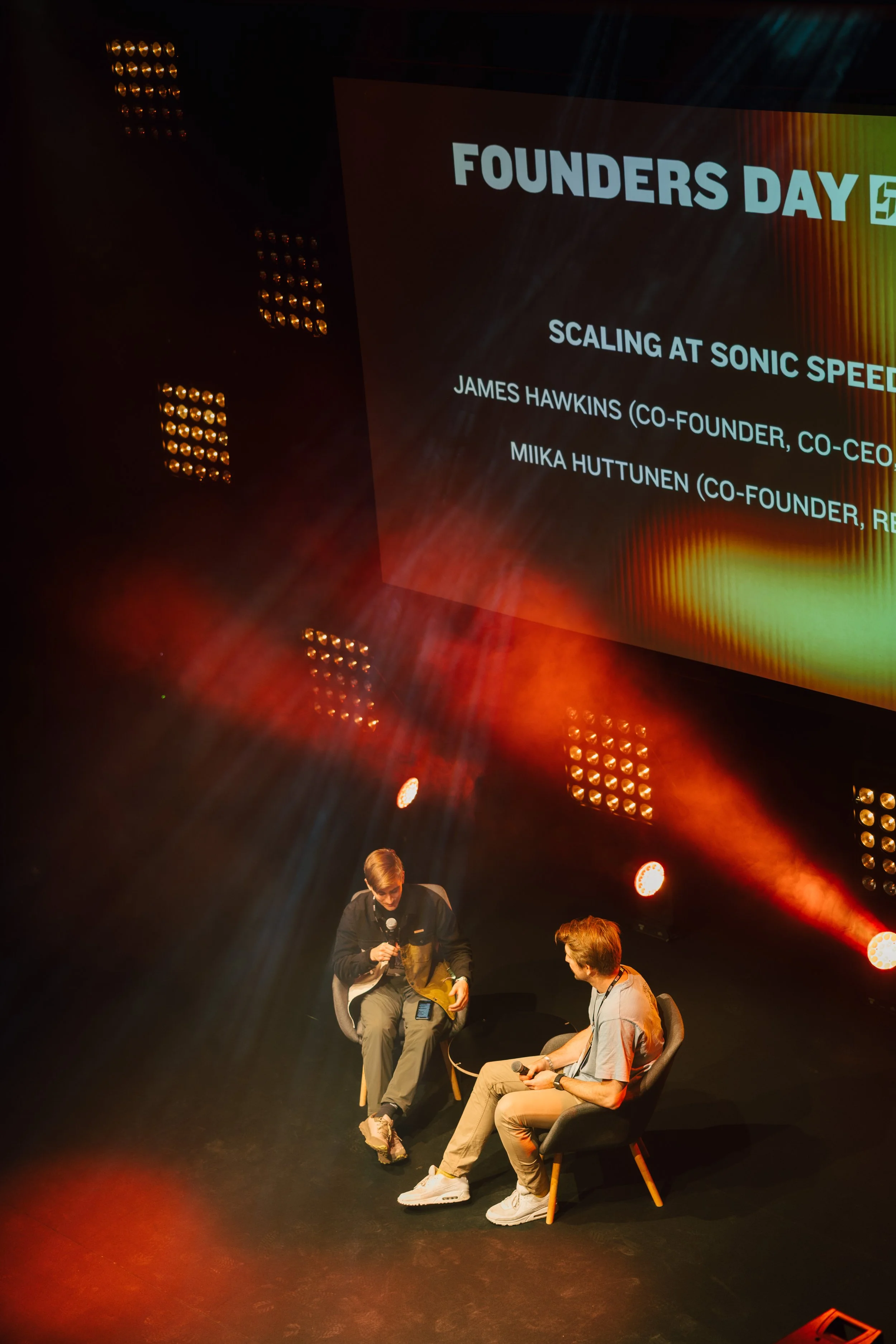 Two men sitting on stage in discussion during a conference. A large screen behind them displays text about Founders Day and scaling at Sonic Speed, with names James Hawkins and Miika Huttunen.