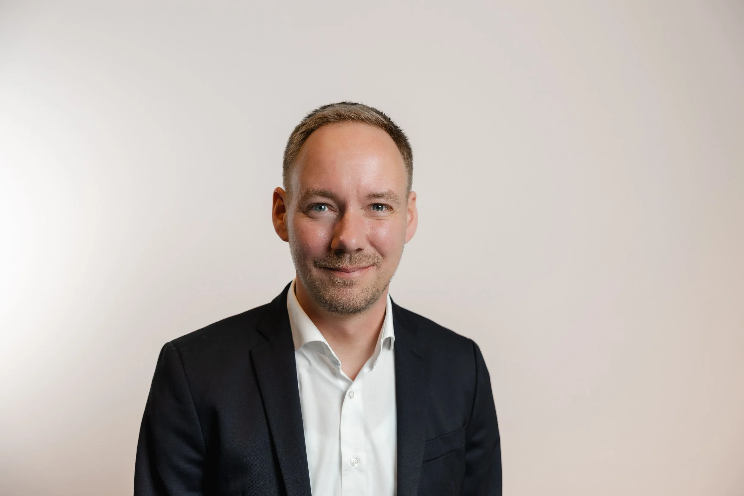 Professional headshot of a man in a black suit and white shirt, smiling slightly against a plain white background.