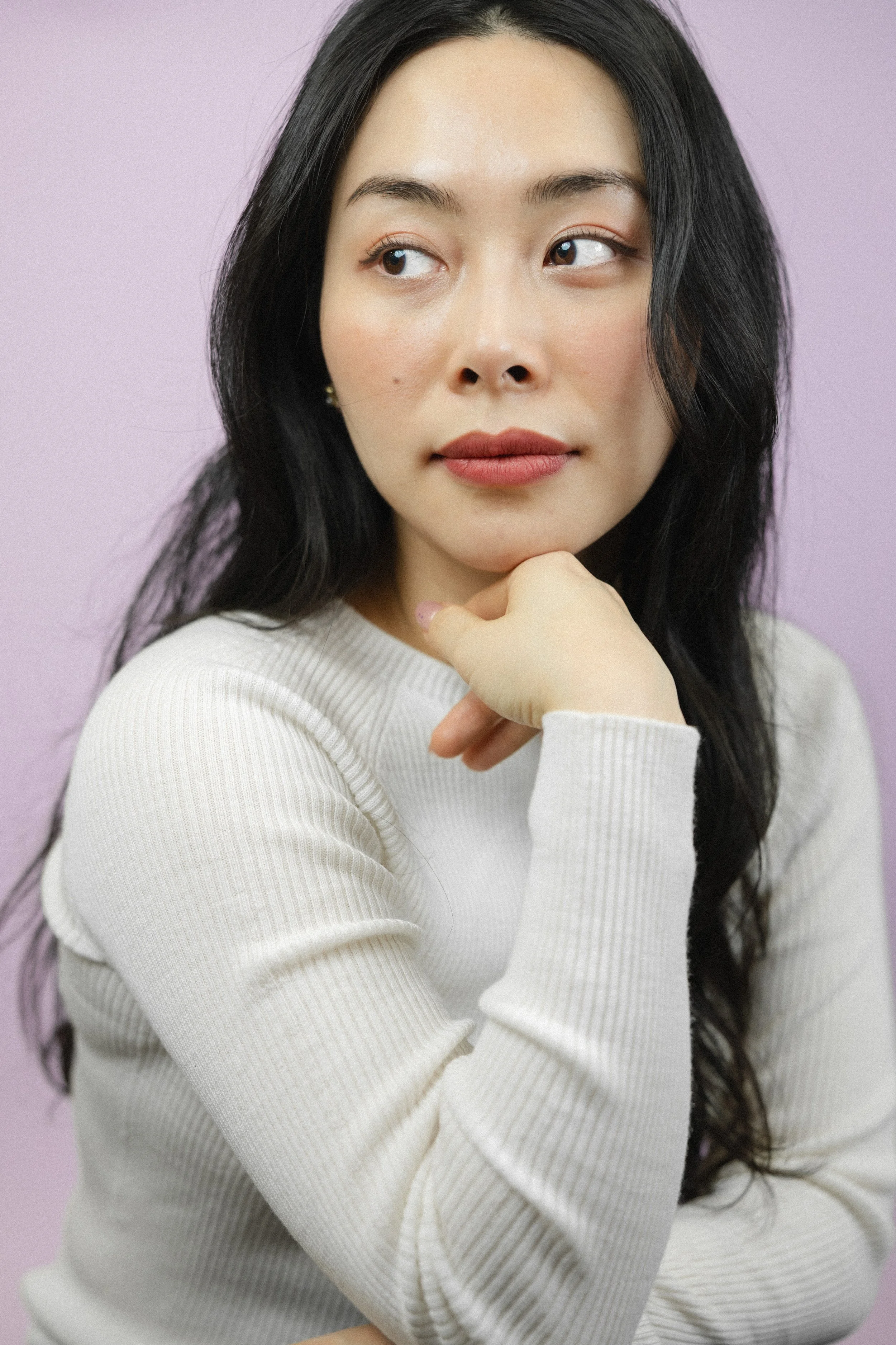 A portrait of a woman with long black hair, light skin, and wearing a white ribbed sweater, against a soft purple background. She is resting her chin on her hand and looking to the side.