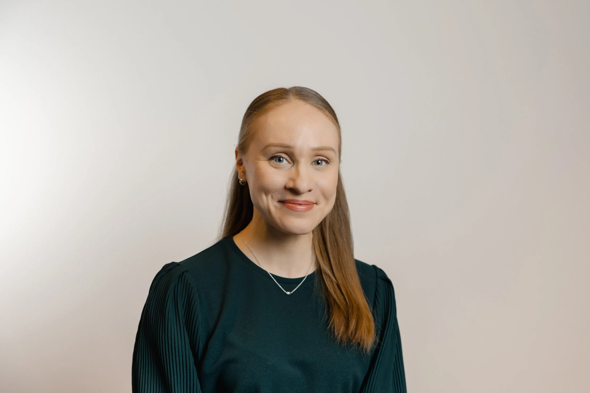 A woman with long red hair, blue eyes, and light skin, smiling softly and wearing a black top and jewelry, standing against a plain white background.