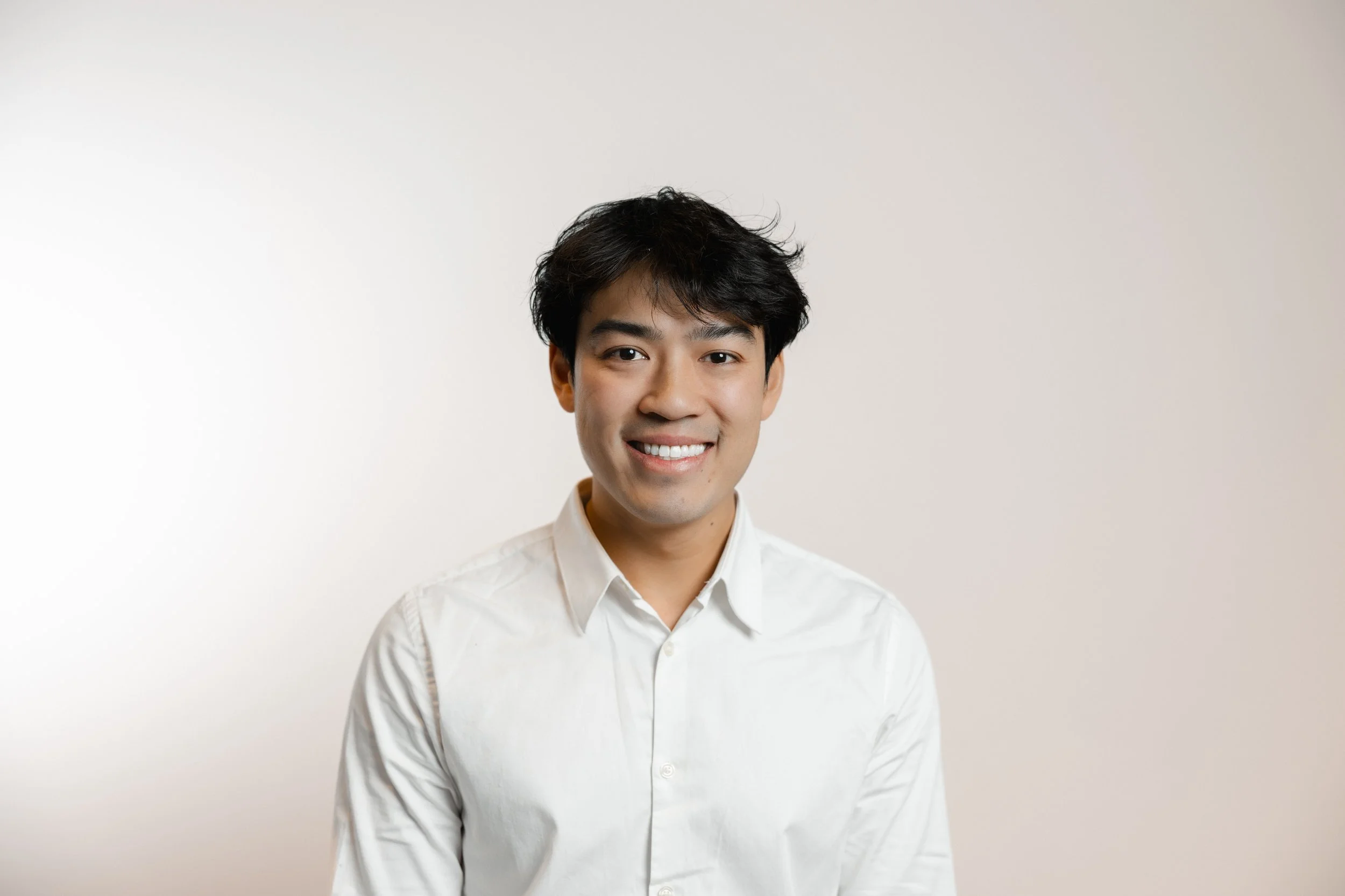 Young man with black hair and a white button-up shirt smiling against a plain off-white background.