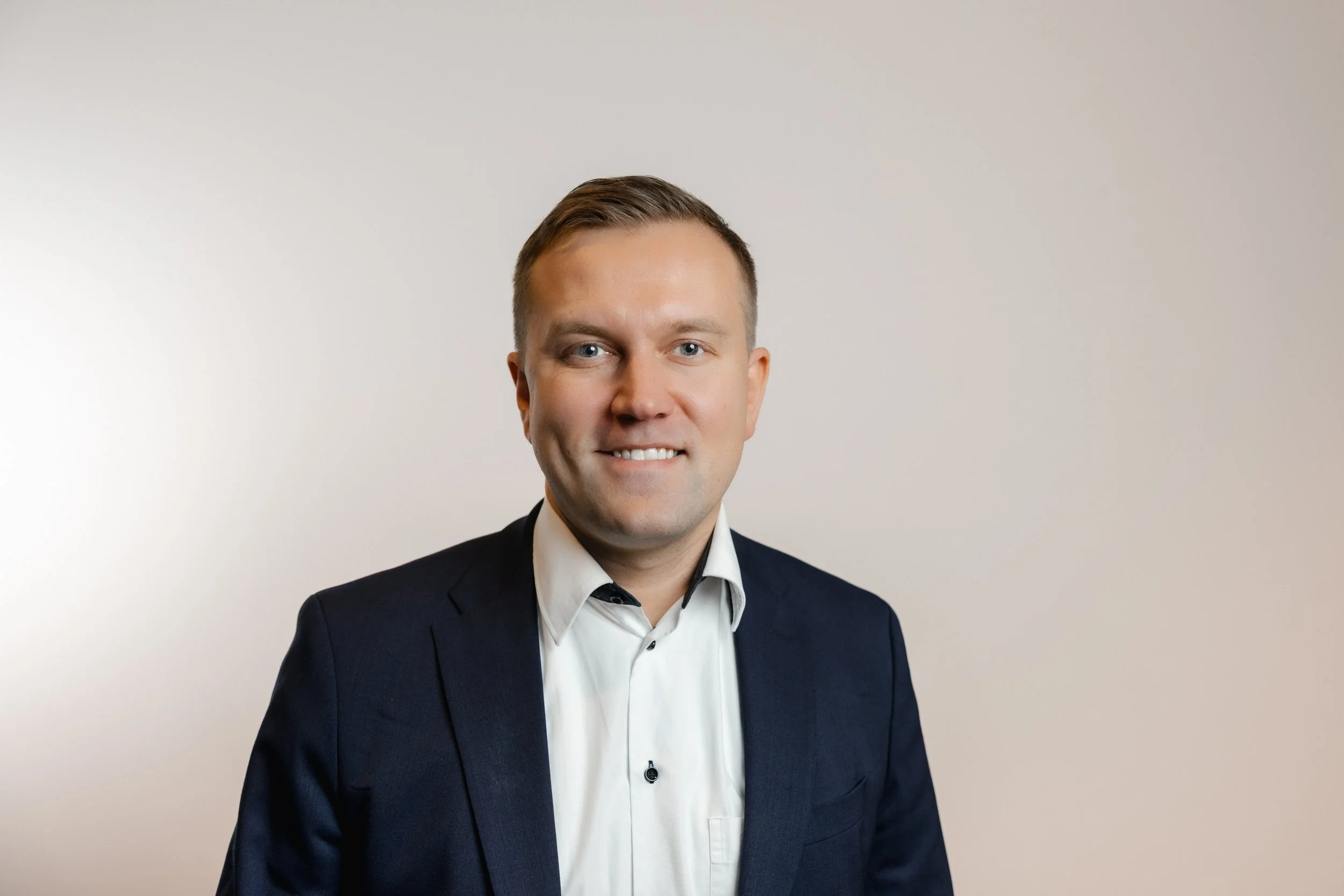 A man in a dark suit and white shirt smiling against a plain light-colored background.