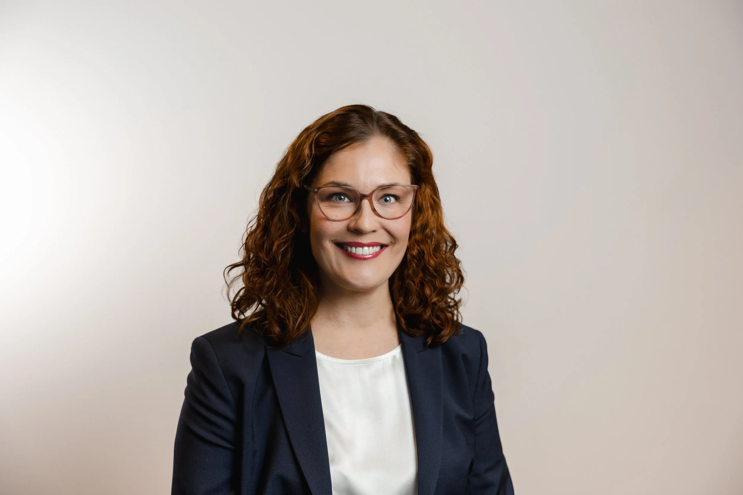 A smiling woman with curly red hair, glasses, a dark blazer, and a white top against a plain light background.