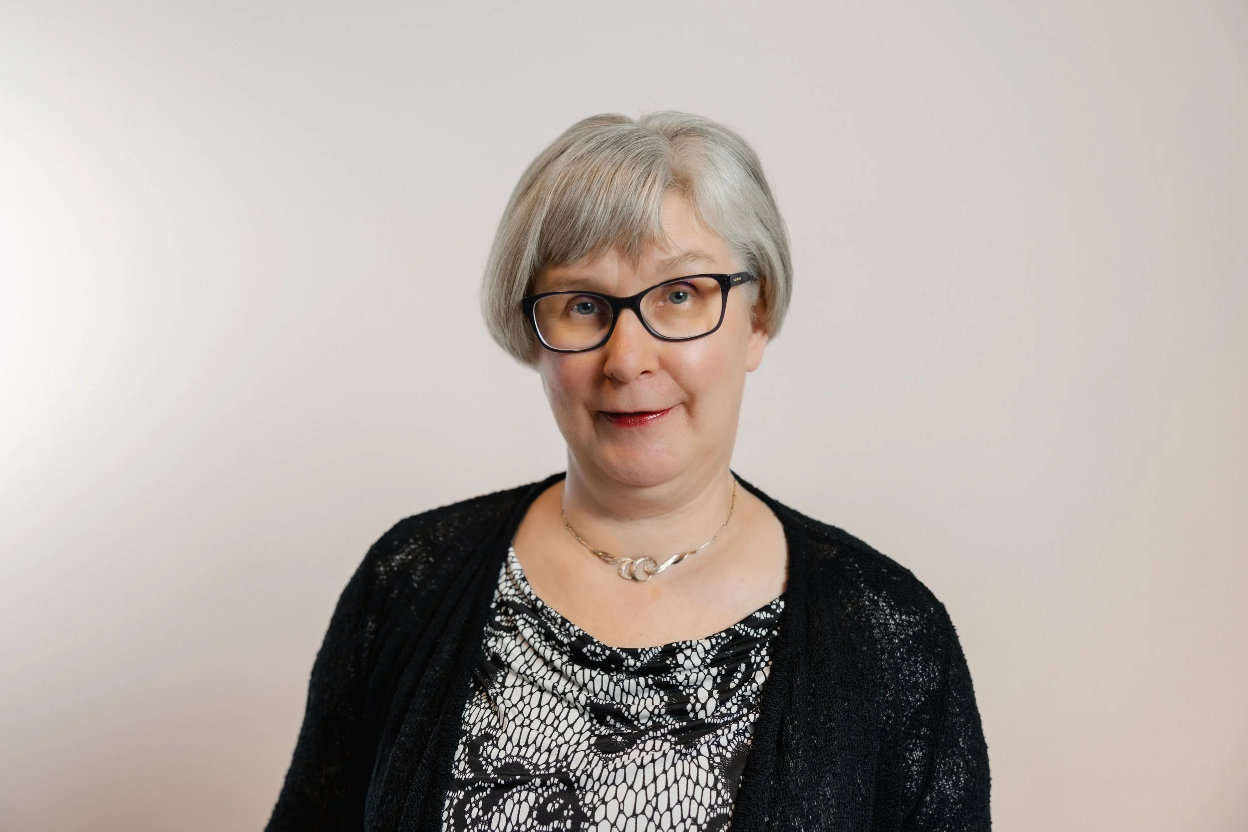 A woman with short gray hair, wearing glasses, a black lace cardigan, a patterned blouse, and a silver necklace, standing against a plain beige background.