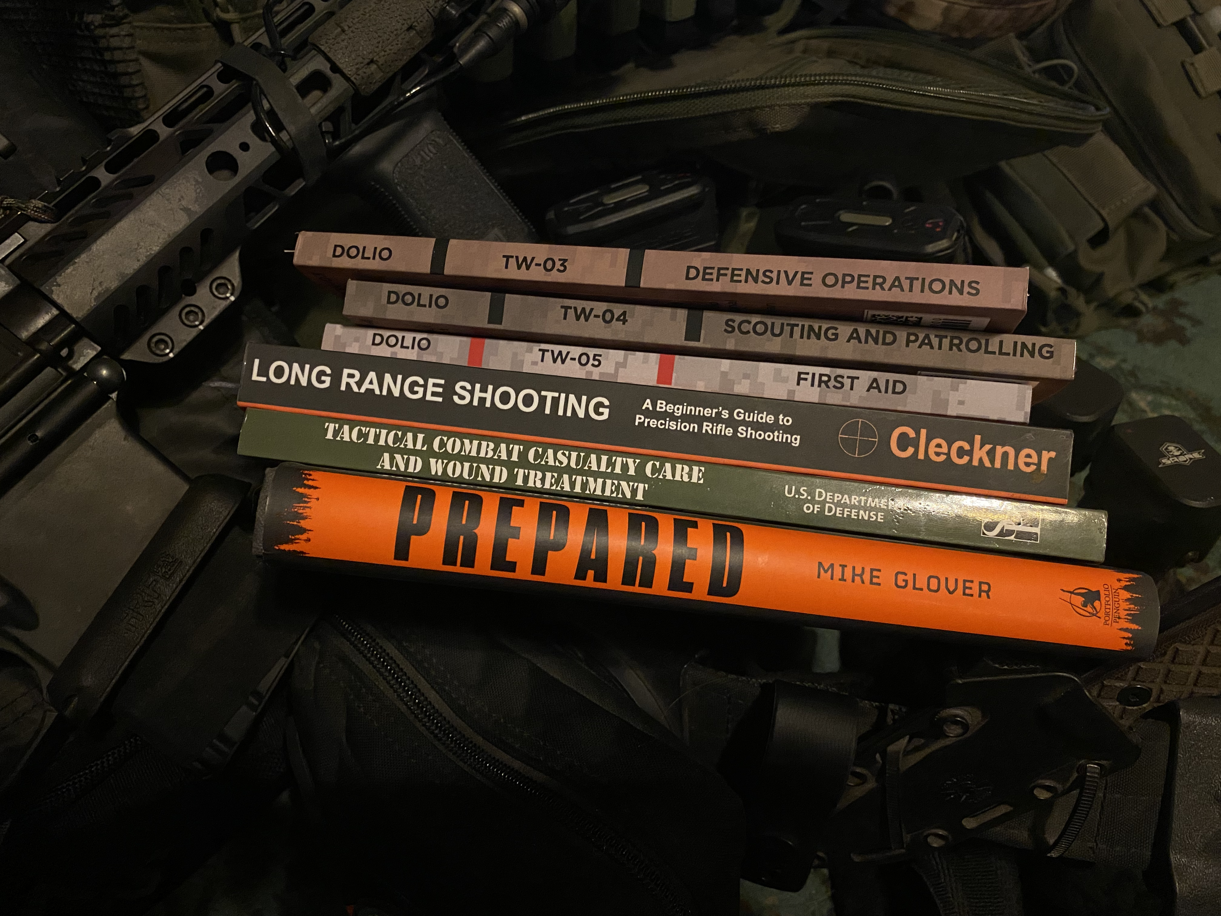Stack of books on top of training gear. Dolio TW-03 Defensive Operations. Dolio TW-04 Scouting and Patrolling. TW-05 First Aid. Long Range Shooting Cleckner. Tactical Combat Casualty Care and Wound Treatment. Prepared Mike Glover.