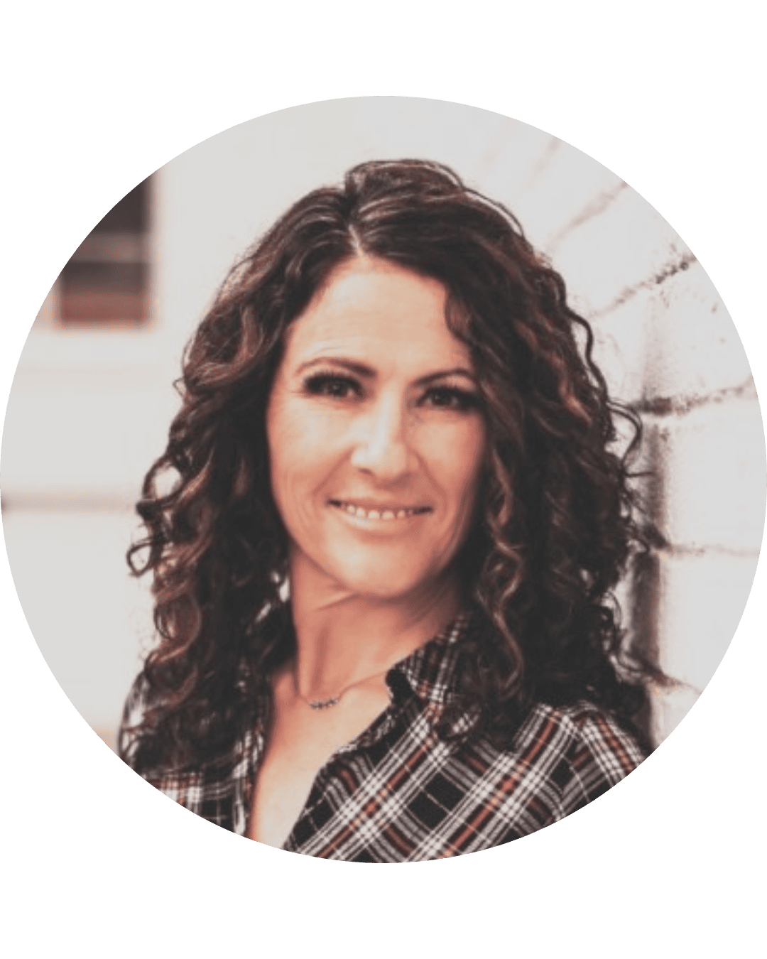 A woman with curly brown hair smiling and leaning against a white brick wall.