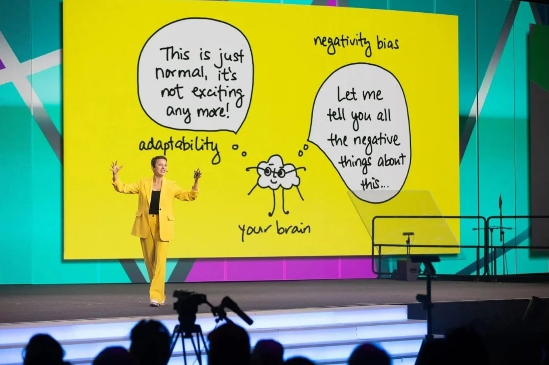A woman in a bright yellow suit giving a presentation on stage. A large yellow poster behind her depicts a cartoon brain with glasses and two speech bubbles. One speech bubble says, 'This is just normal, it's not exciting any more!' and the other says, 'Let me tell you all the negative things about this...' The poster is labeled 'negativity bias' and 'adaptability.' Audience members are visible in the foreground, with cameras pointed at the stage.