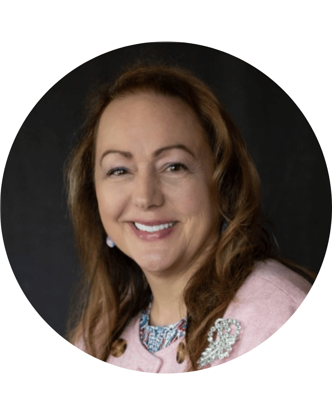 Headshot of a woman smiling, wearing a pink blazer with a jeweled brooch, and a patterned blouse, against a dark background.