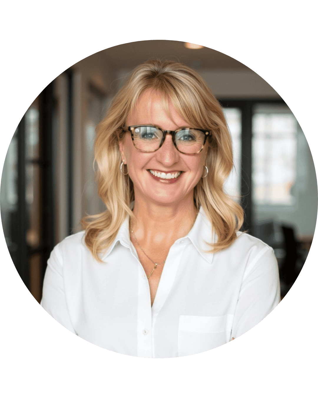 A woman with blonde hair, glasses, and earrings smiling in an office environment, wearing a white shirt.