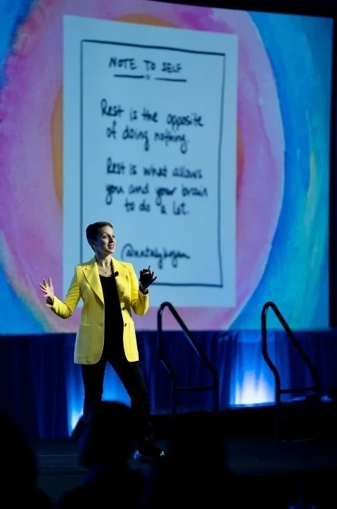 A woman in a yellow blazer giving a presentation on stage. Behind her, a large screen displays a handwritten note with motivational advice, including a quote about self-care.