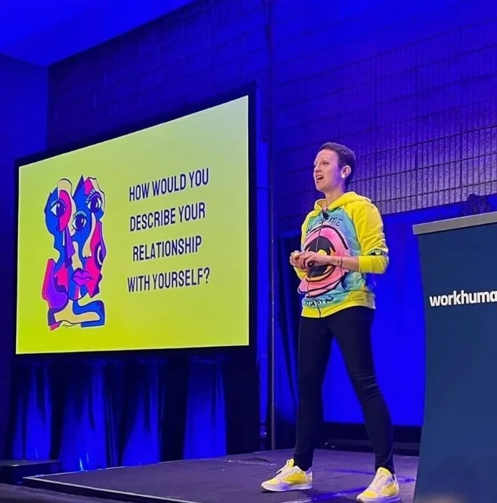 Presentation slide with a colorful abstract face illustration and the text "How would you describe your relationship with yourself?" A woman in a vibrant hoodie and yellow sneakers stands on stage asking the question.
