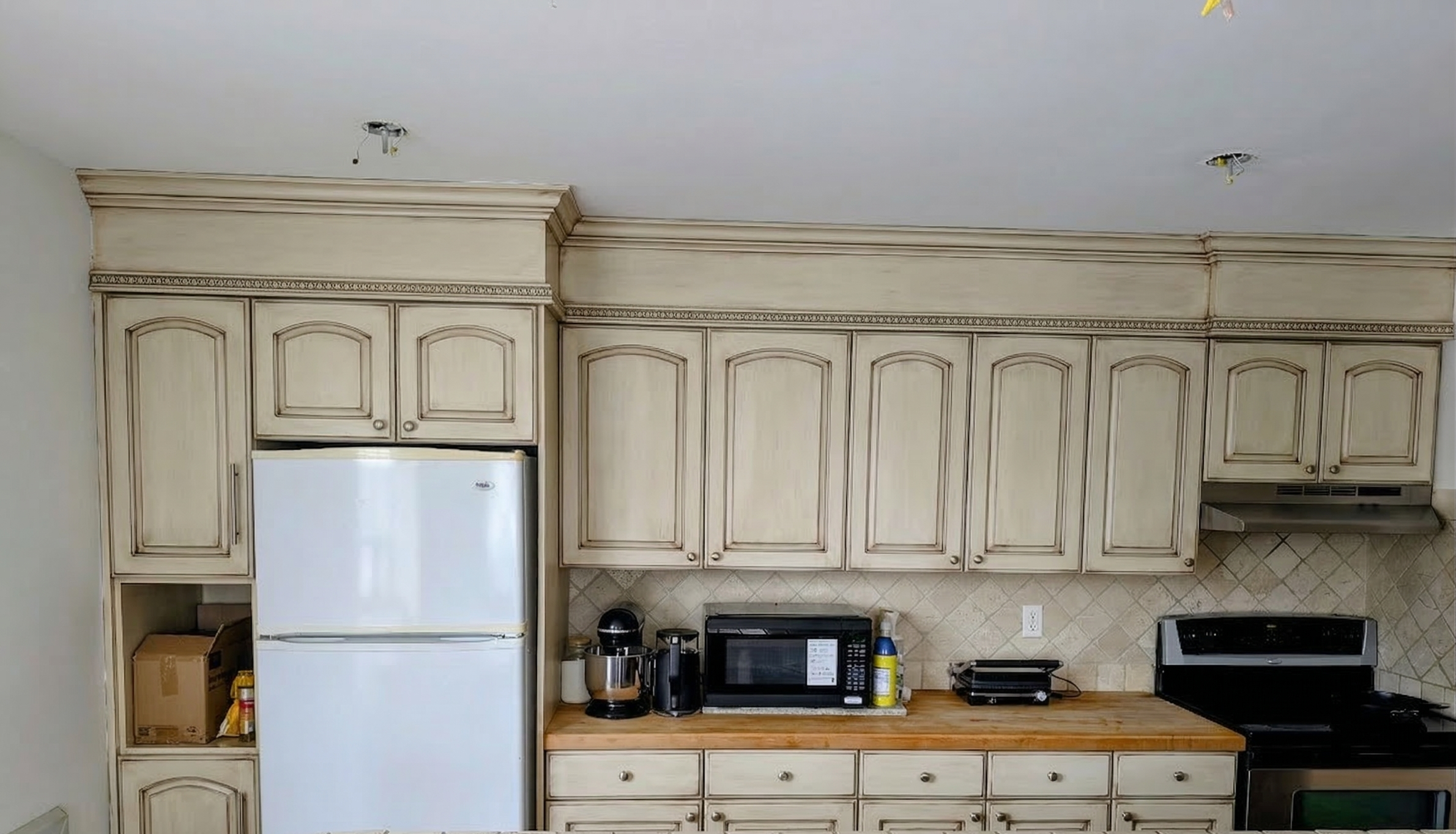Kitchen with white cabinets, wooden countertop, white refrigerator, microwave, coffee maker, toaster, and stove, with neutral tile backsplash.
