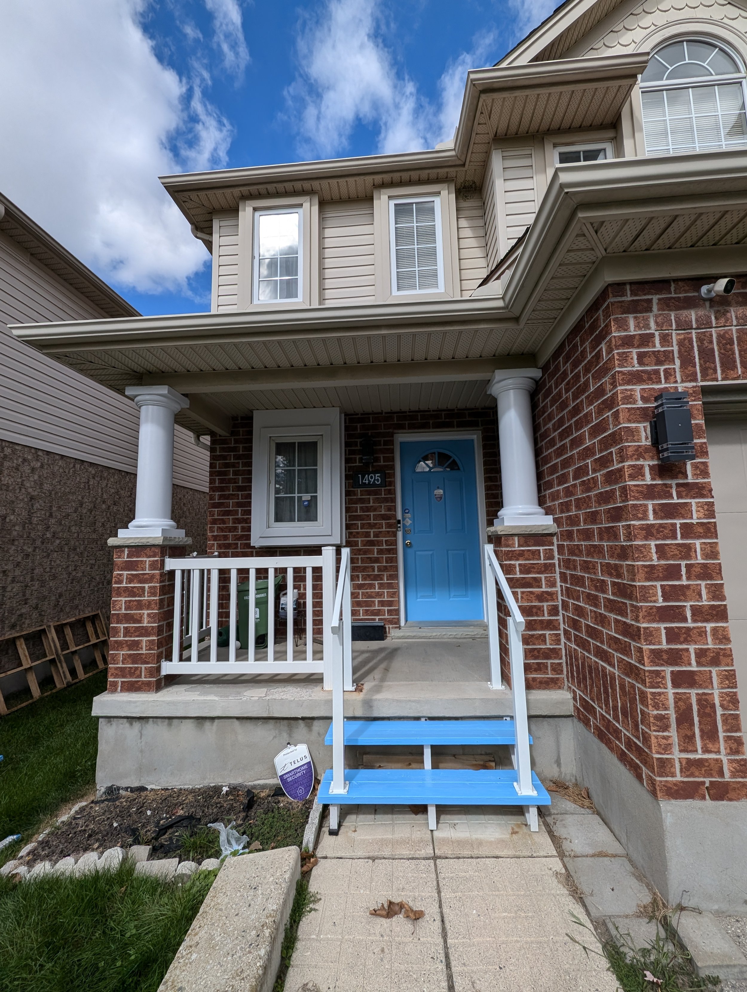 A two-story house with a brick facade, blue front door, small front porch with white railing, and a blue staircase leading up to the porch. The house has beige siding, white trim around windows, and two large columns supporting the porch roof. There 