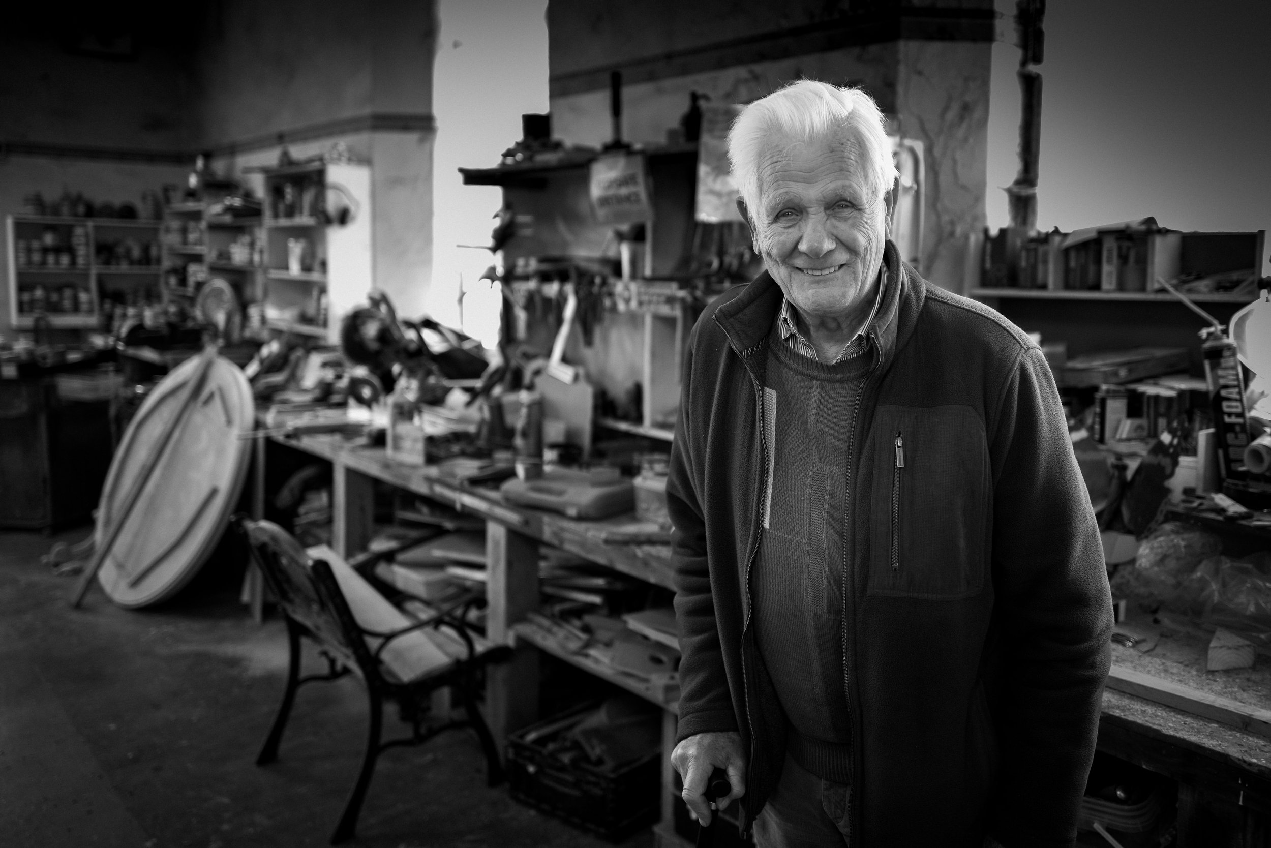 An elderly man smiling in a workshop or cluttered space with shelves and tools in the background