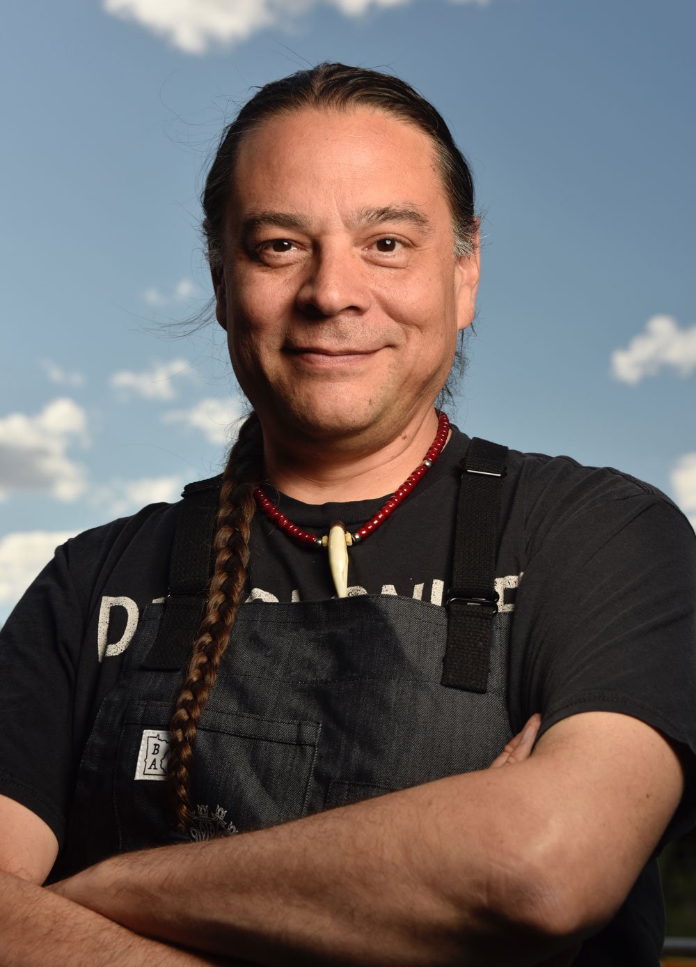Sean Sherman has light skin, his arms are crossed and he is wearing a grey apron with a black t-shirt underneath; the background is a blue sky.