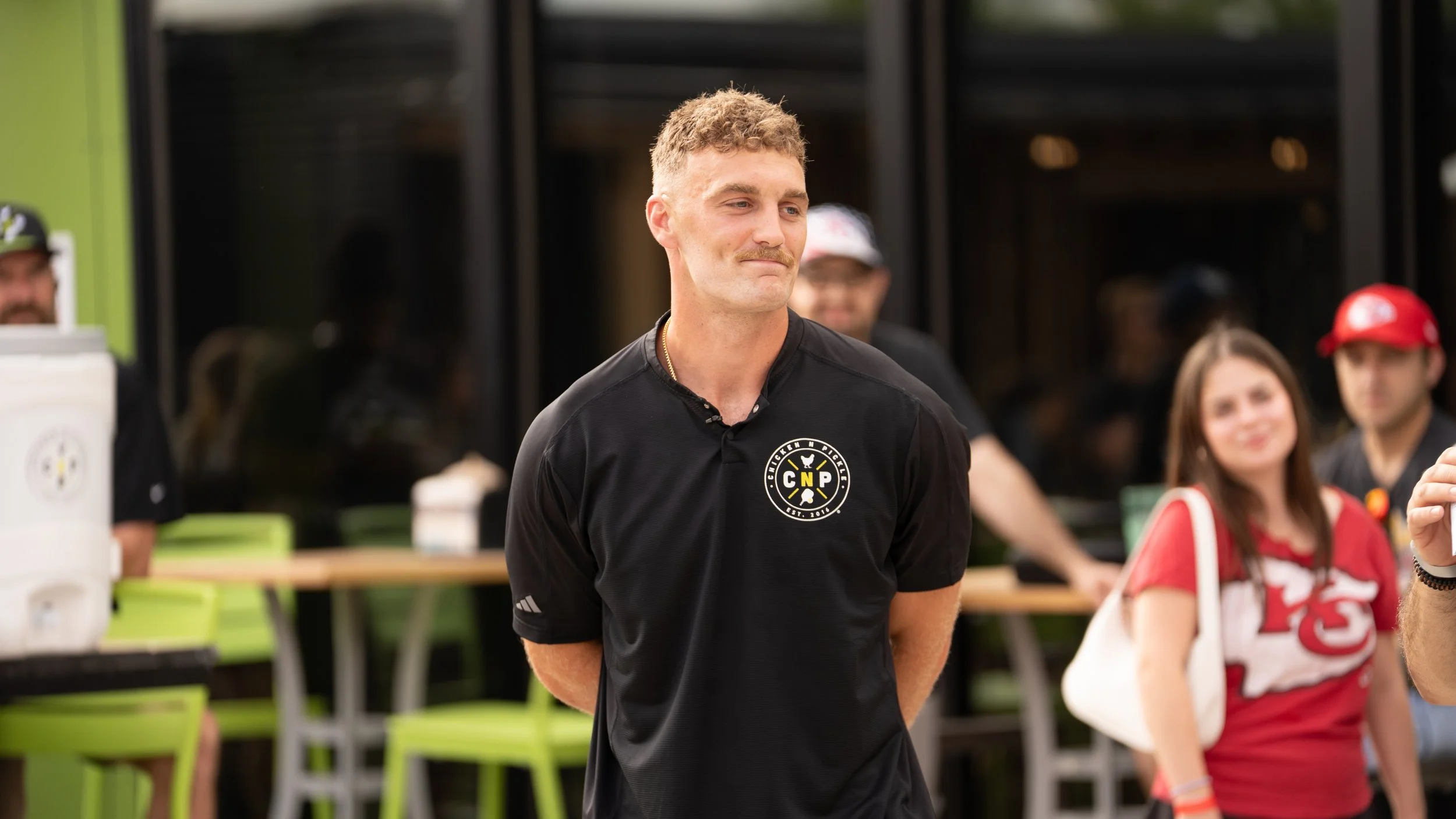 A young man with short, curly hair, wearing a black shirt with a logo, stands outdoors with a slight smile, surrounded by people.