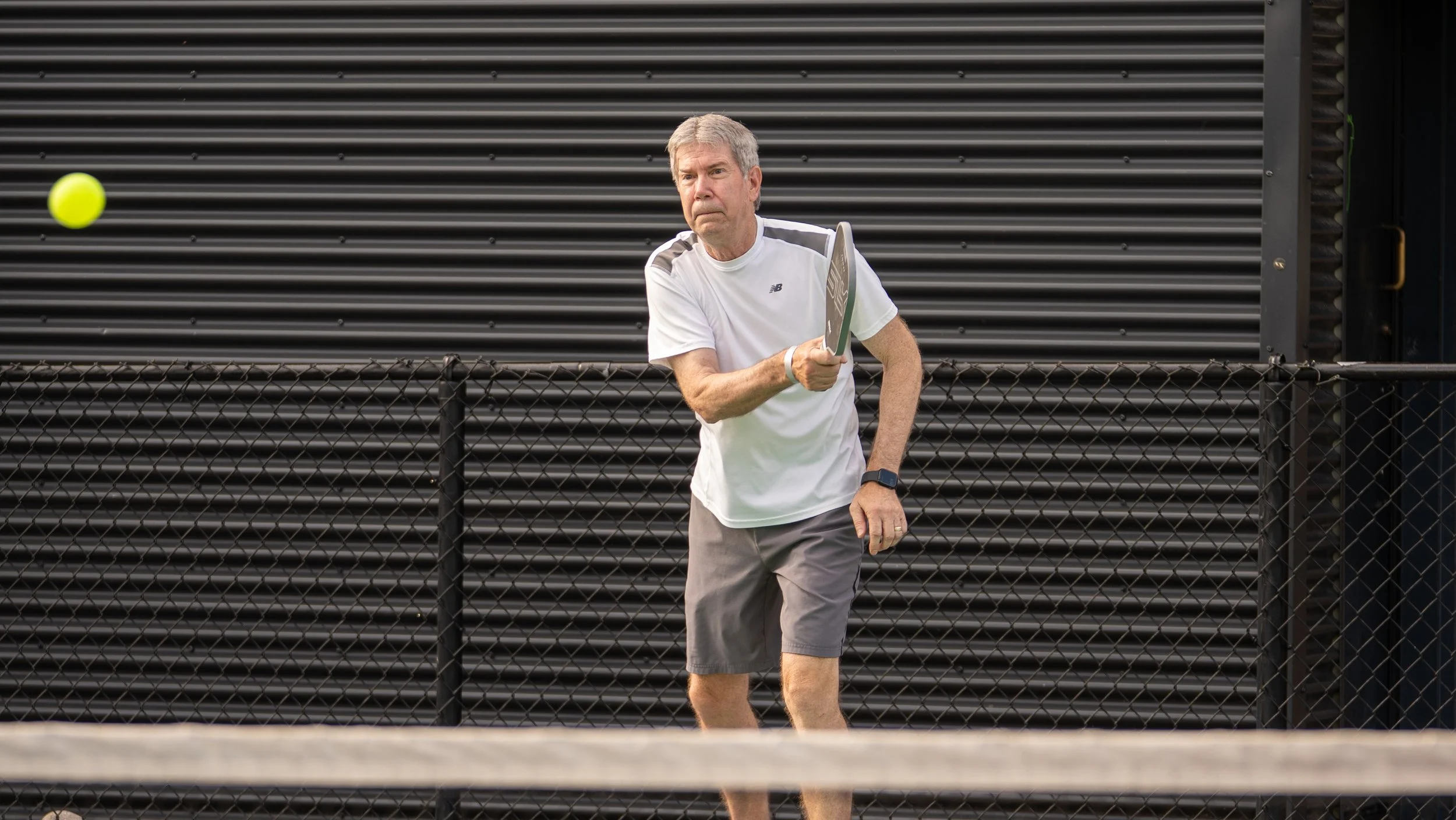 Older man playing pickleball, holding a paddle, in front of a black metal fence and building.