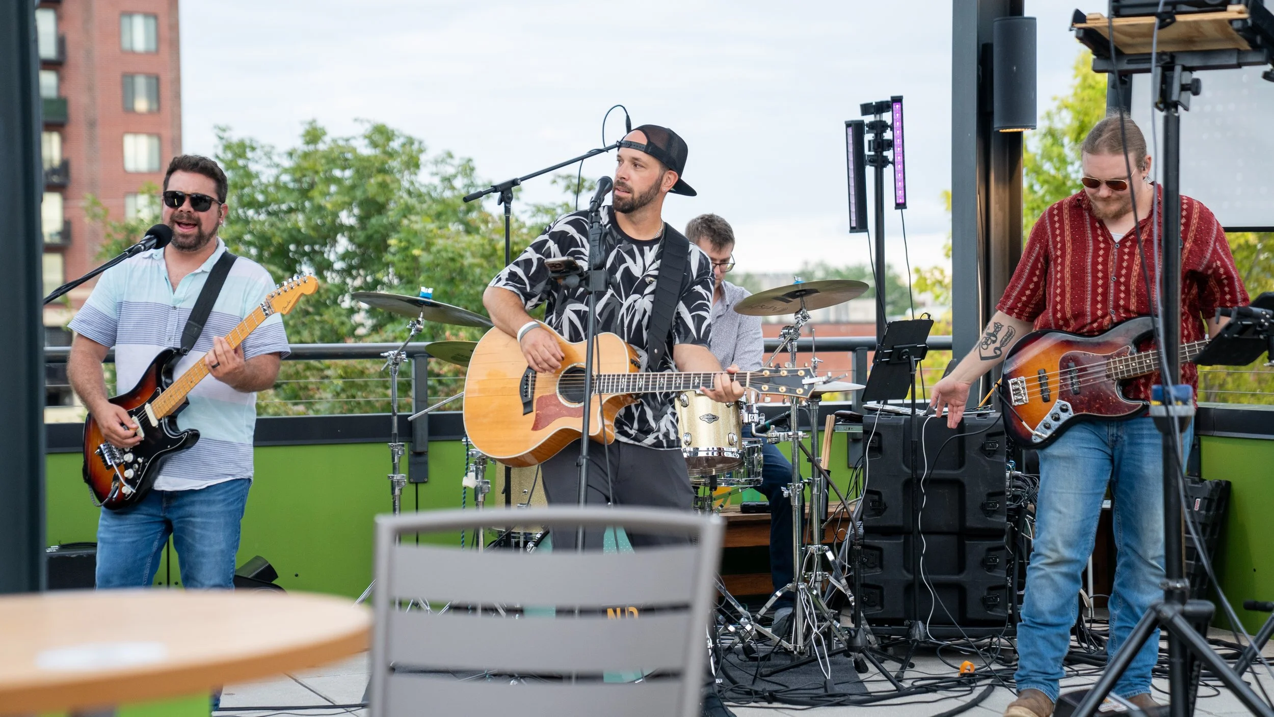 A band of four musicians performing on an outdoor stage, with one playing guitar, another on drums, and two on bass guitars, with greenery and buildings in the background.