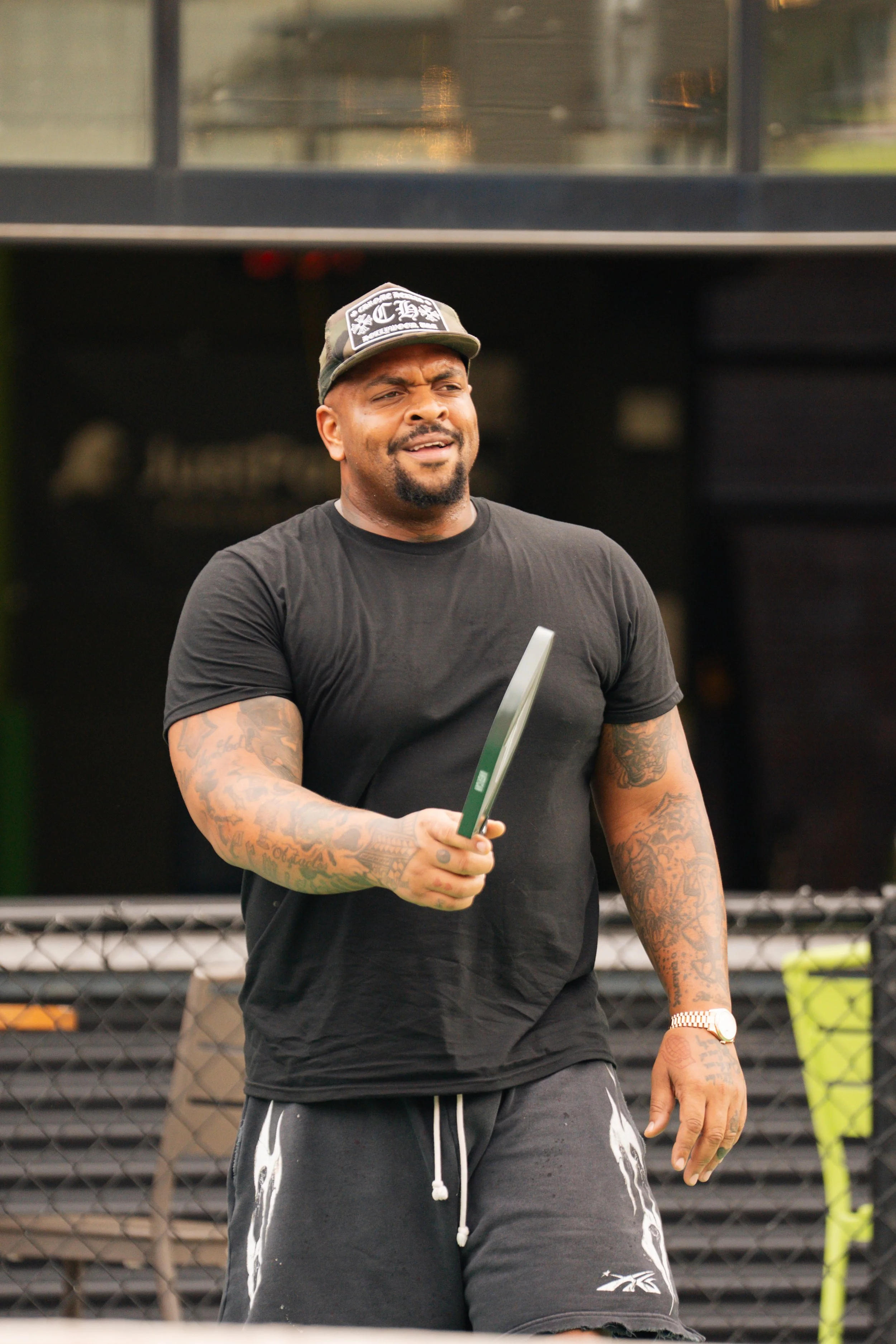 Man in black t-shirt and shorts holding a tablet, with tattoos on both arms, standing outdoors.