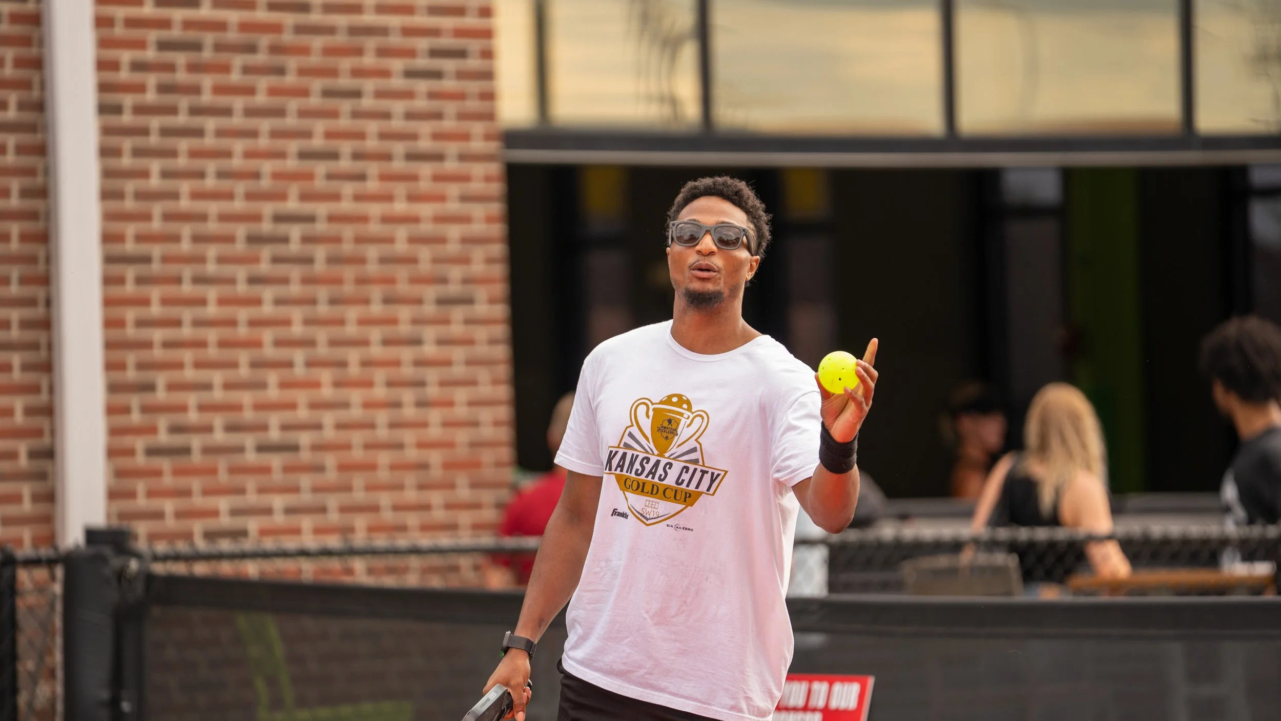A young man with sunglasses holding a yellow tennis ball and a tennis racket, standing outdoors near a brick building, with people in the background.