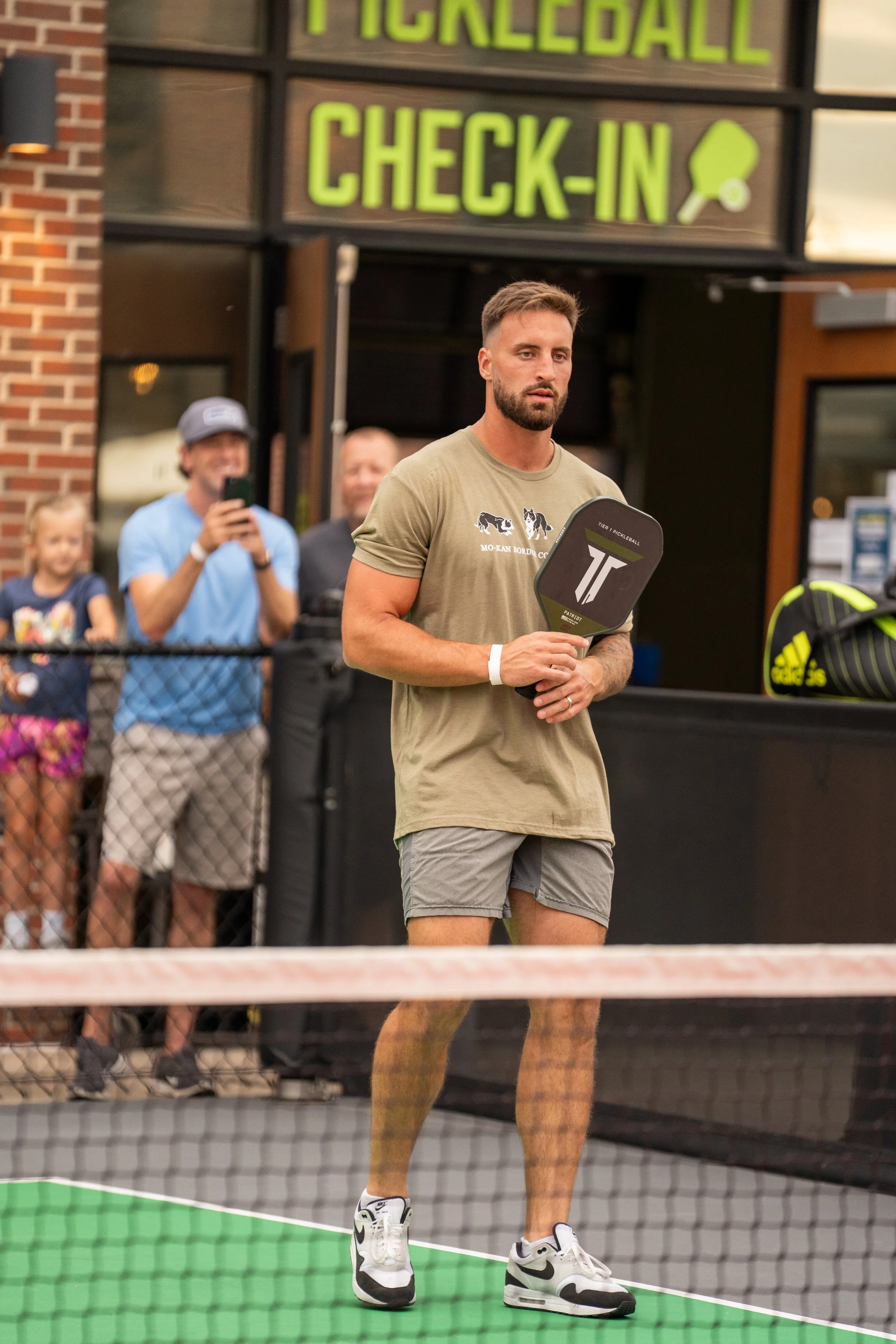 A man holding a pickleball paddle standing on a pickleball court with people watching in the background, including a woman with a phone, outside a building with a sign that reads 'PICKLEBALL CHECK-IN'.