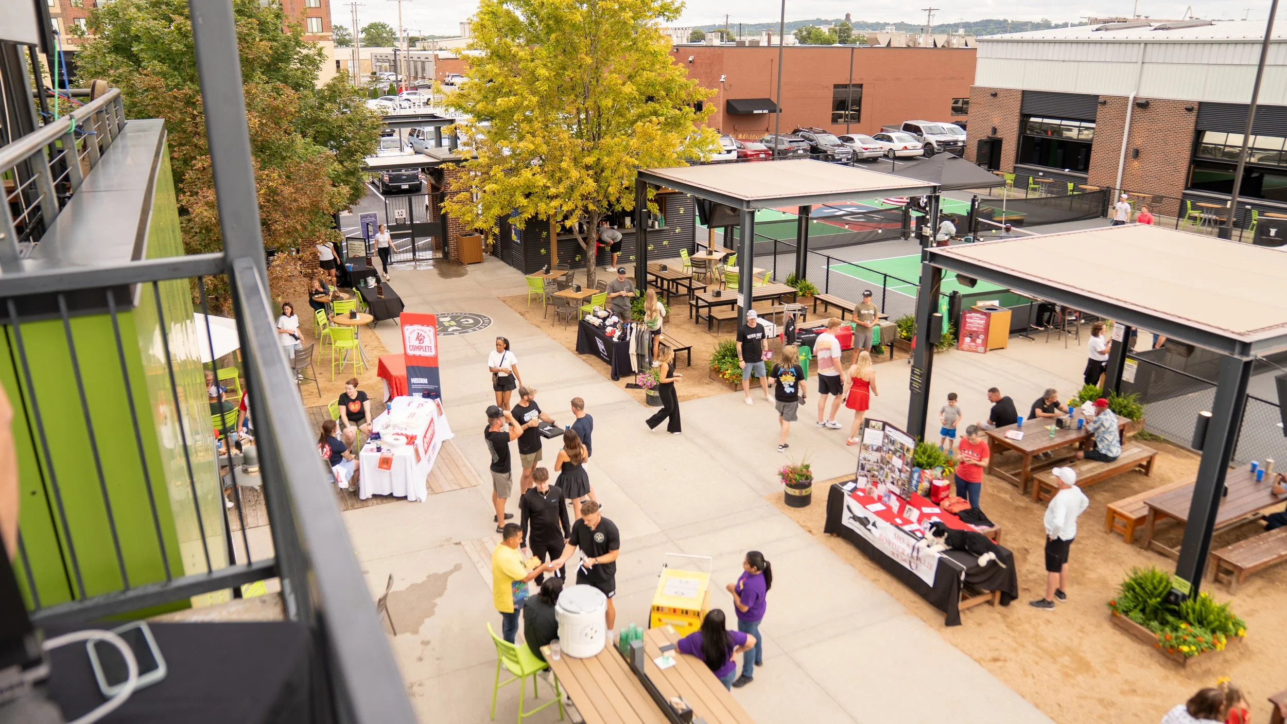 Outdoor event with various booths, tables, and seating areas, with people walking, sitting, and interacting. A tree provides shade, and there are tennis courts in the background.