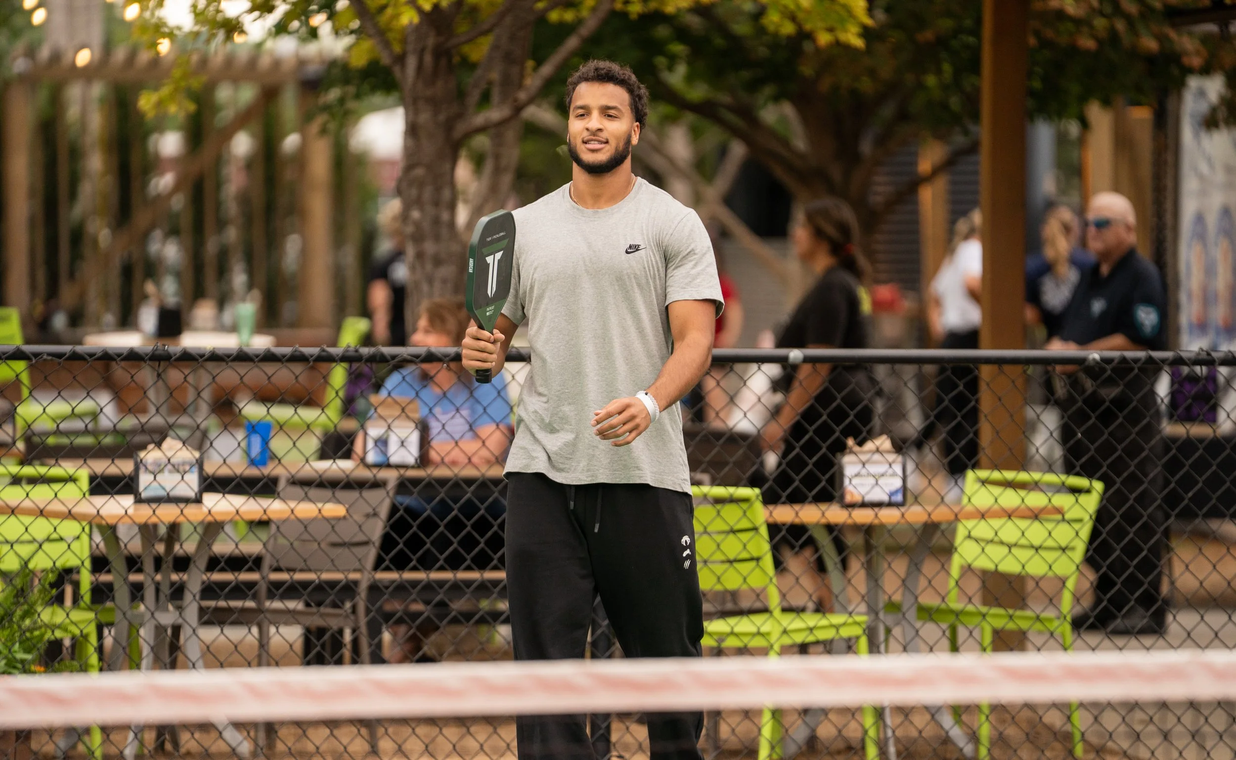 A young man standing outdoors holding a pickleball paddle, dressed in a grey Nike t-shirt and black athletic pants, with a chain-link fence and people in the background.