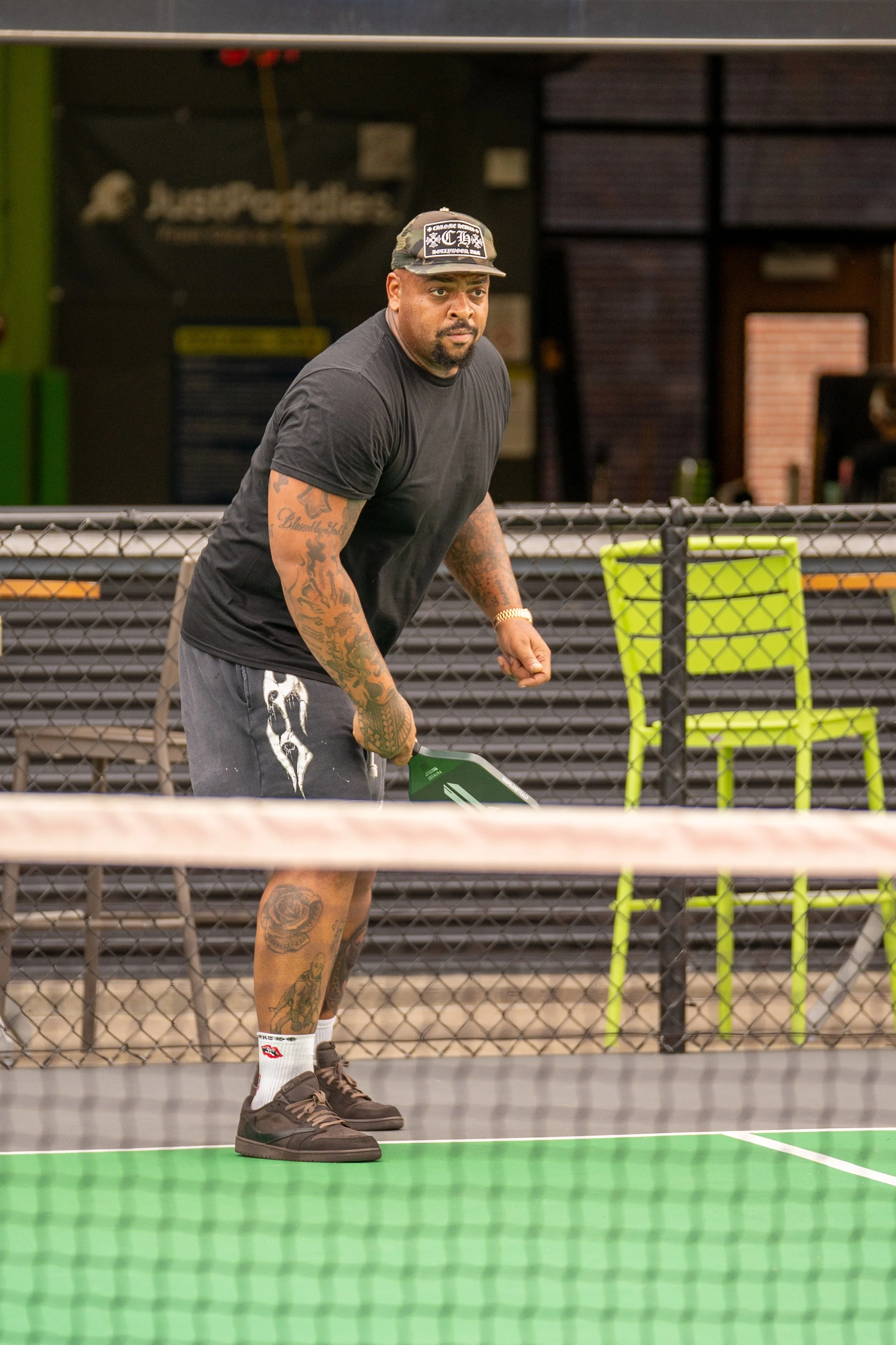 A man with tattoos on his arms and legs playing pickleball on an indoor court, holding a green paddle, with a focused expression.